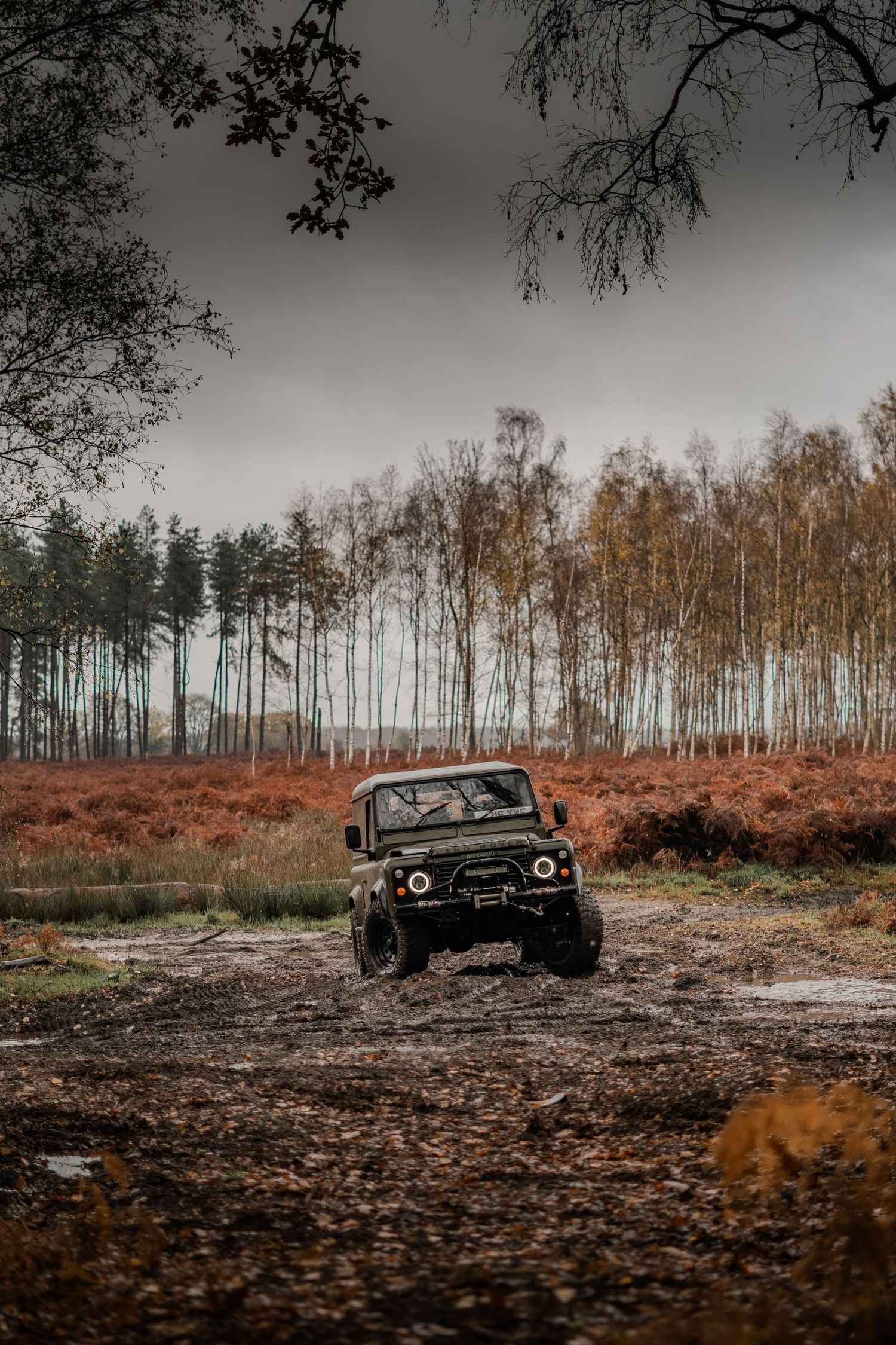 Dark green Land Rover Defender 90 navigating a muddy off-road track surrounded by autumn foliage and birch trees.