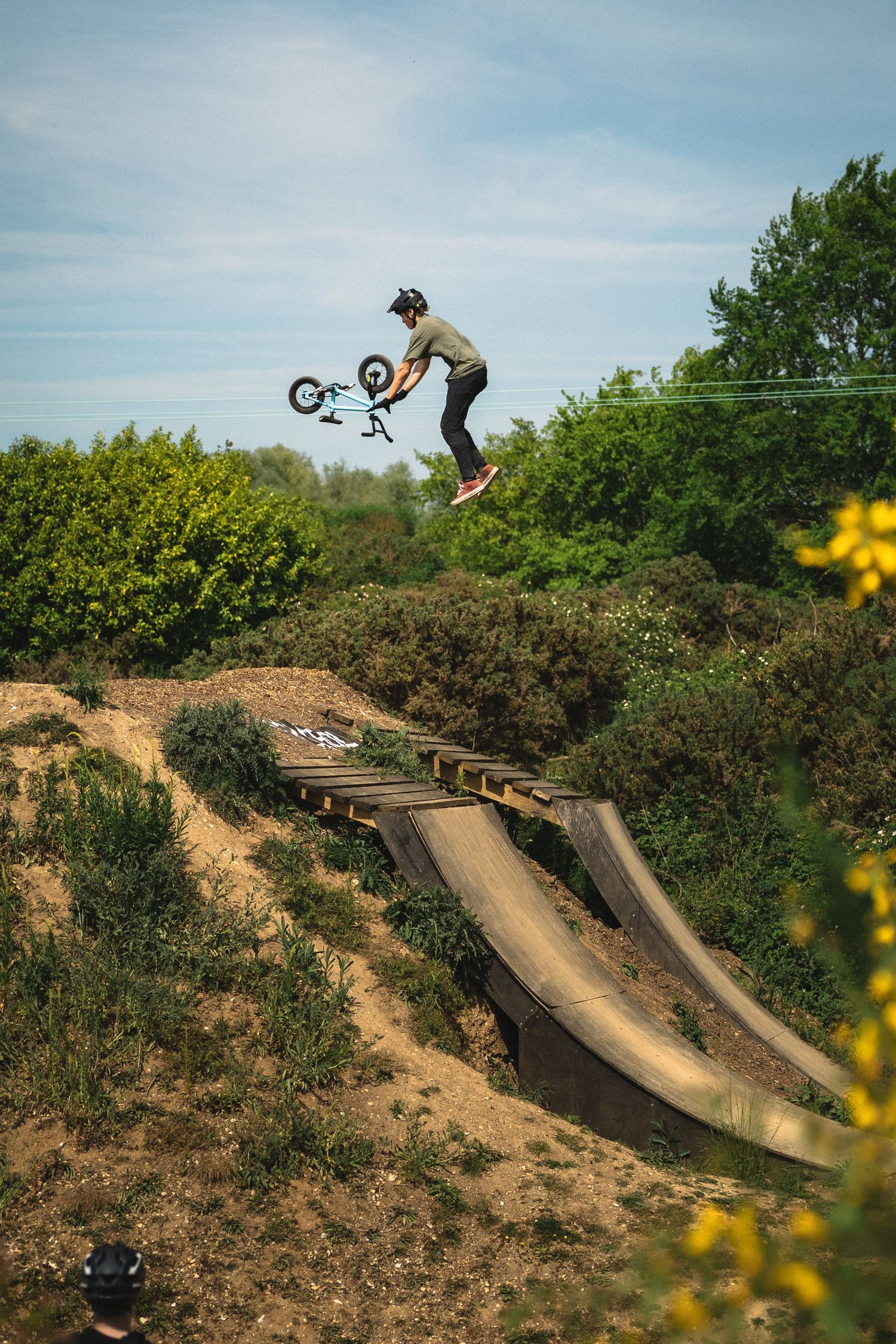 BMX rider performs a trick off a wooden ramp surrounded by green bushes under a blue sky.
