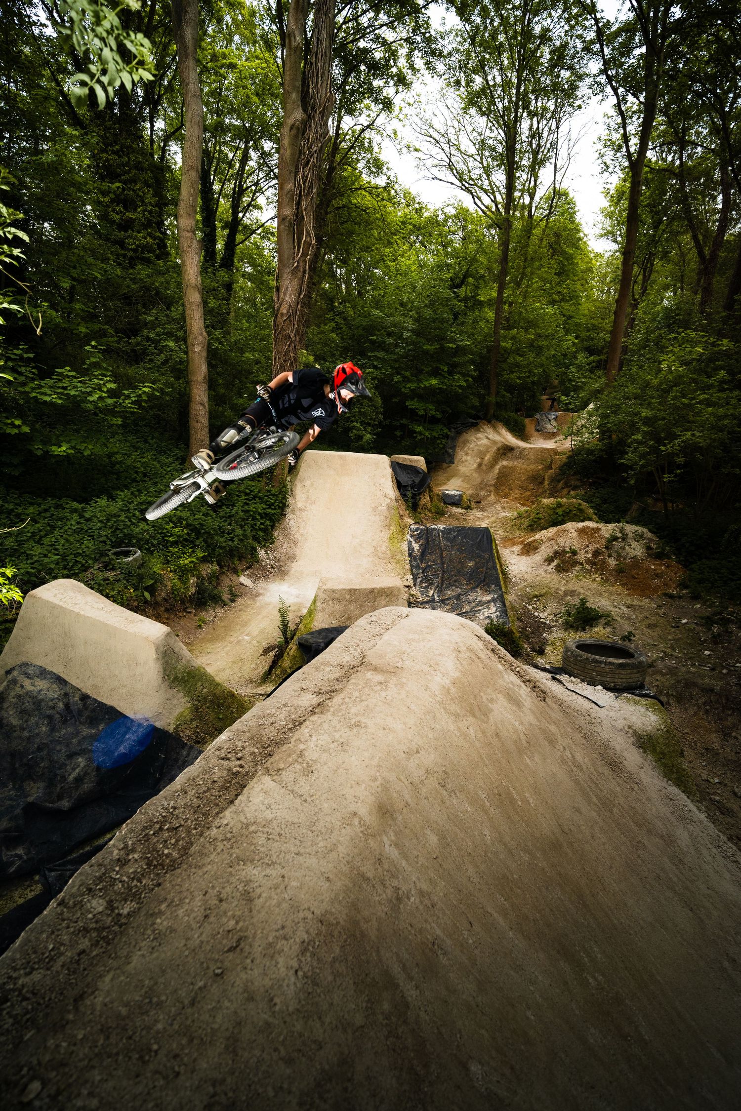 A mountain biker in a red helmet launches off a dirt jump ramp surrounded by lush green forest trees.