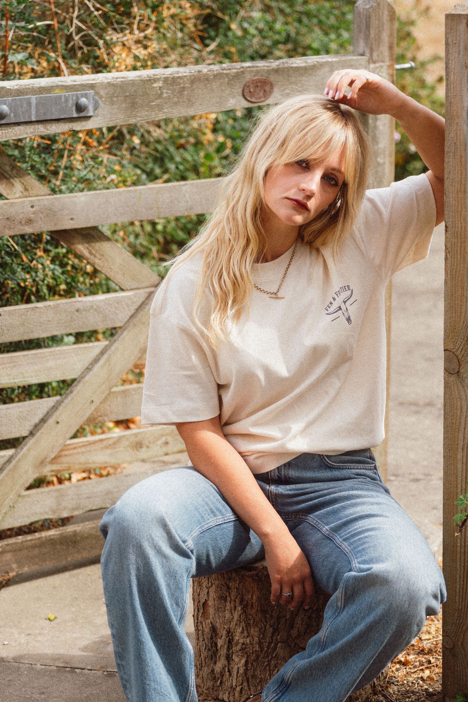 Blonde woman in cream graphic tee and wide-leg jeans sitting on a stump by a wooden fence outdoors.