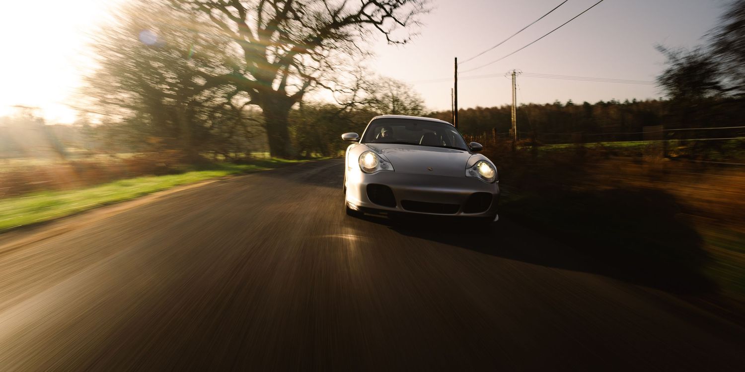 Silver Porsche 911 driving along a tree-lined country road bathed in warm golden sunlight.