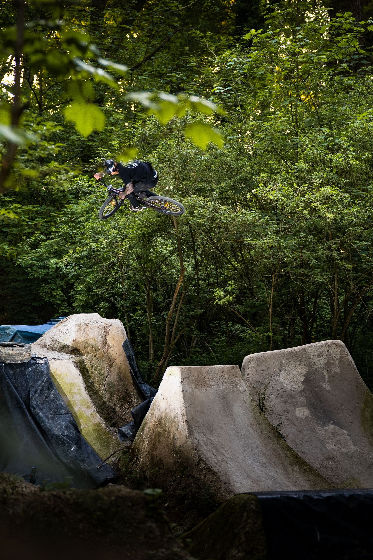BMX rider performing a high aerial trick off concrete ramps in a forest bike park surrounded by lush green trees.
