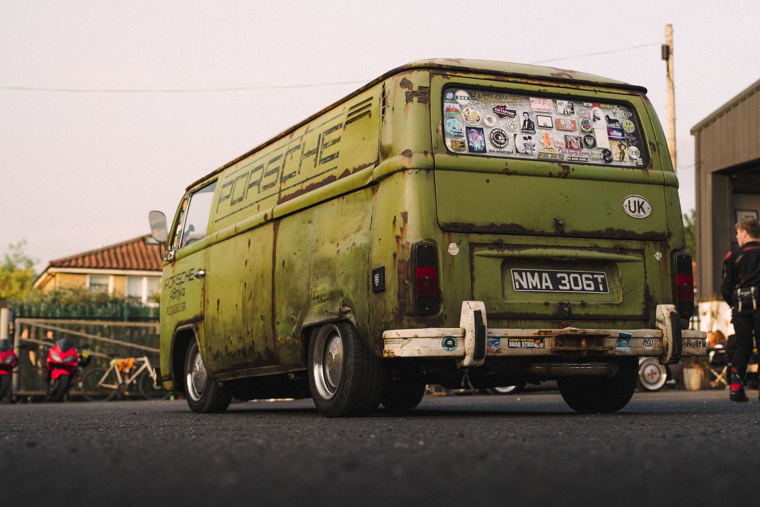 Vintage green Porsche-branded VW van with patina finish and stickers, parked on pavement at dusk.