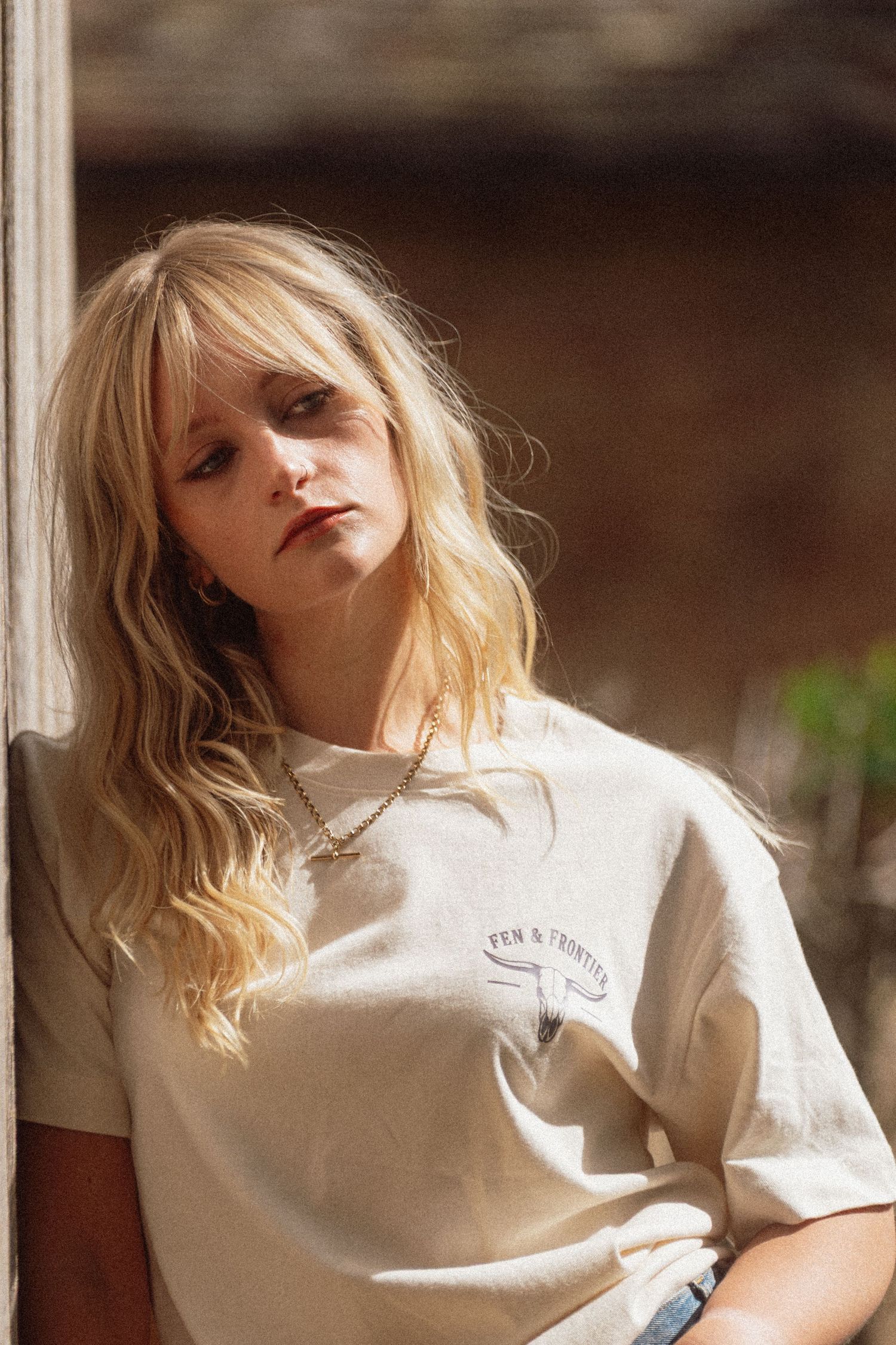 Blonde woman with bangs wearing a beige graphic t-shirt and chain necklace, leaning against a wall in warm sunlight.