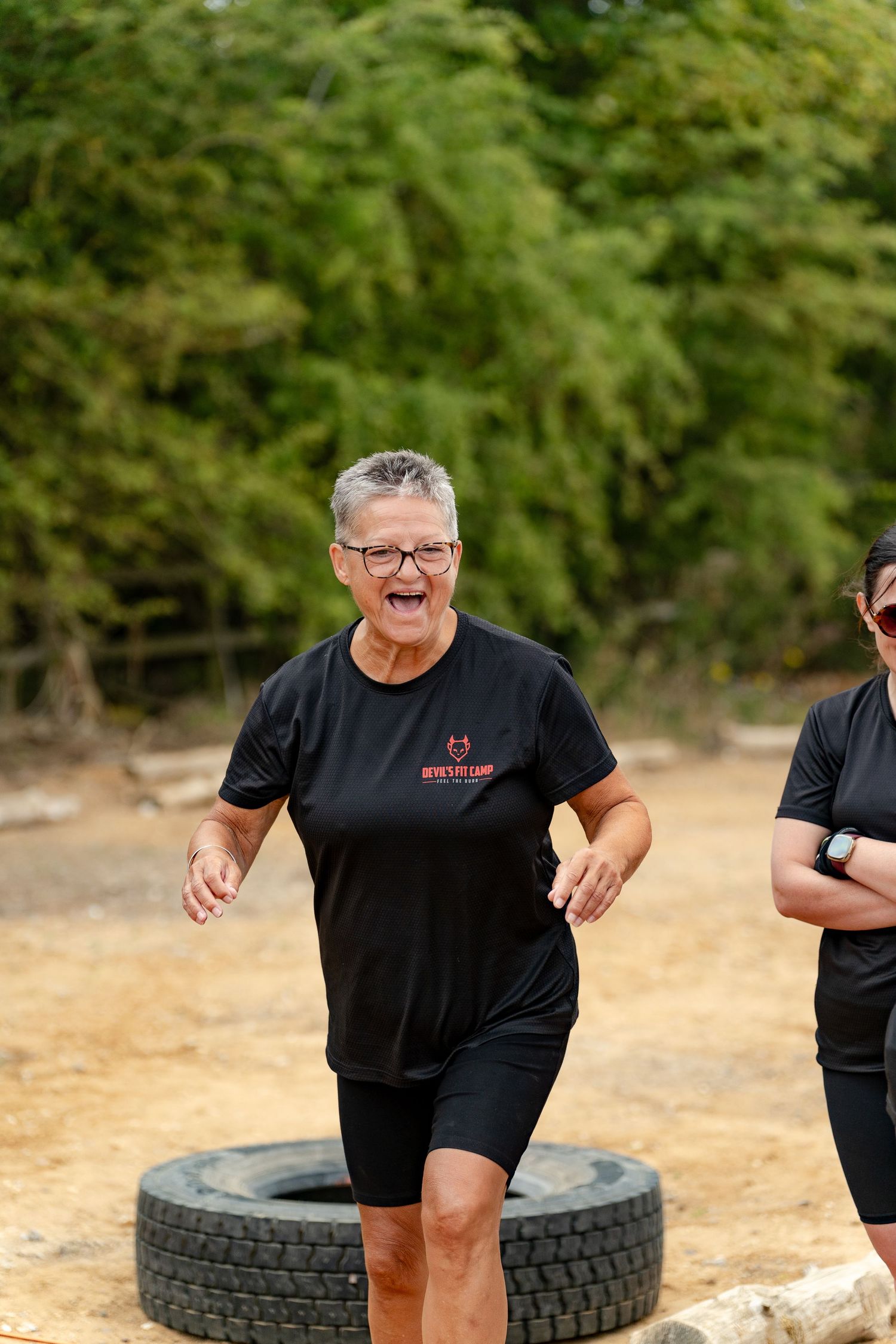 Older woman in black athletic wear laughing while running at an outdoor boot camp fitness course with tires.