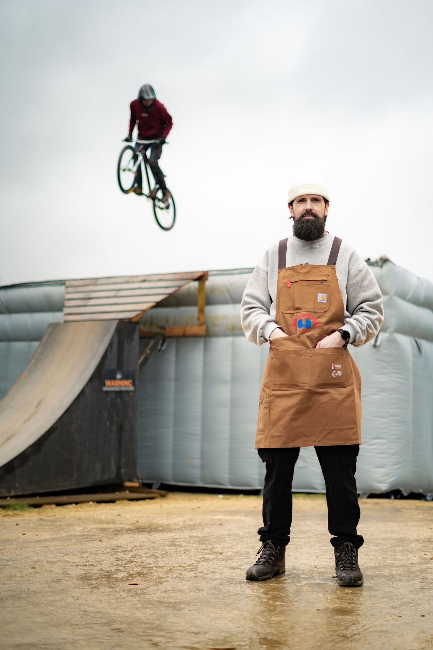 Bearded man in brown apron and hard hat stands at BMX bike park while rider performs aerial trick in background.