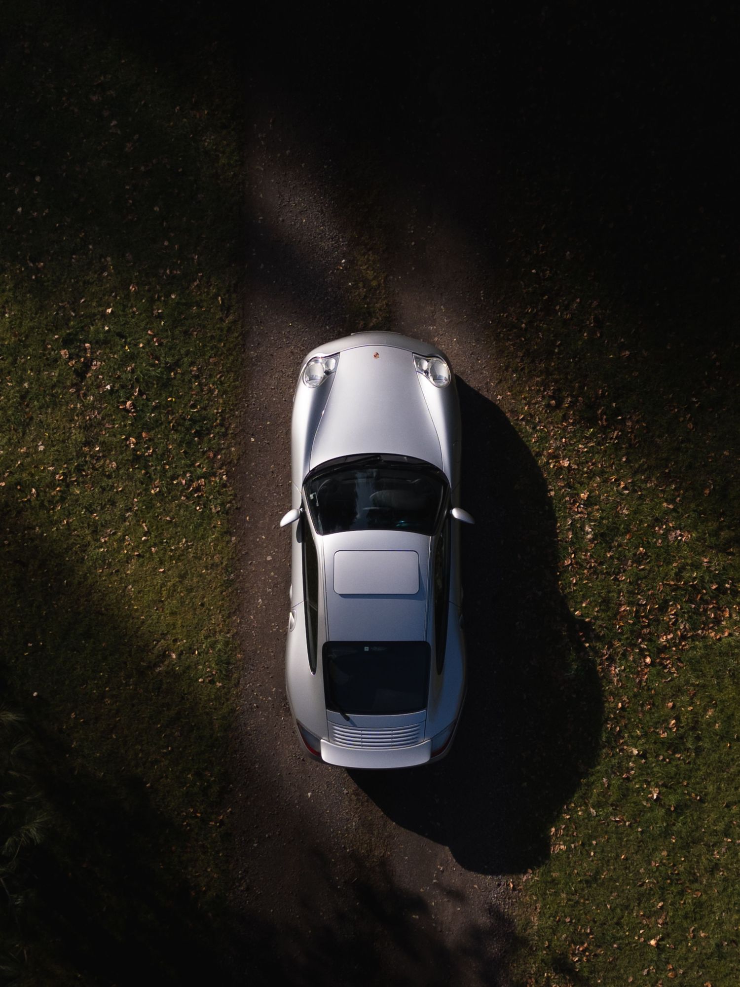 Top-down aerial close-up of a silver Porsche 911 parked on a gravel path bordered by green grass.