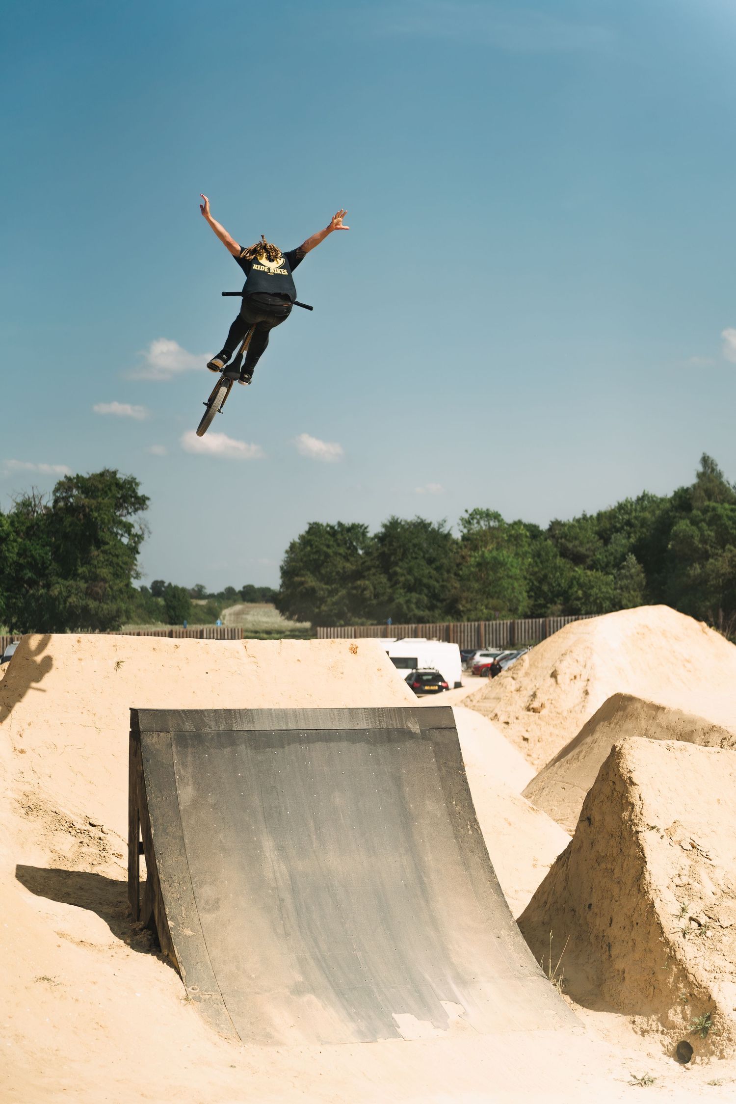 BMX cyclist executing a no-handed aerial jump over a wooden ramp at a sandy outdoor bike park on a sunny day.