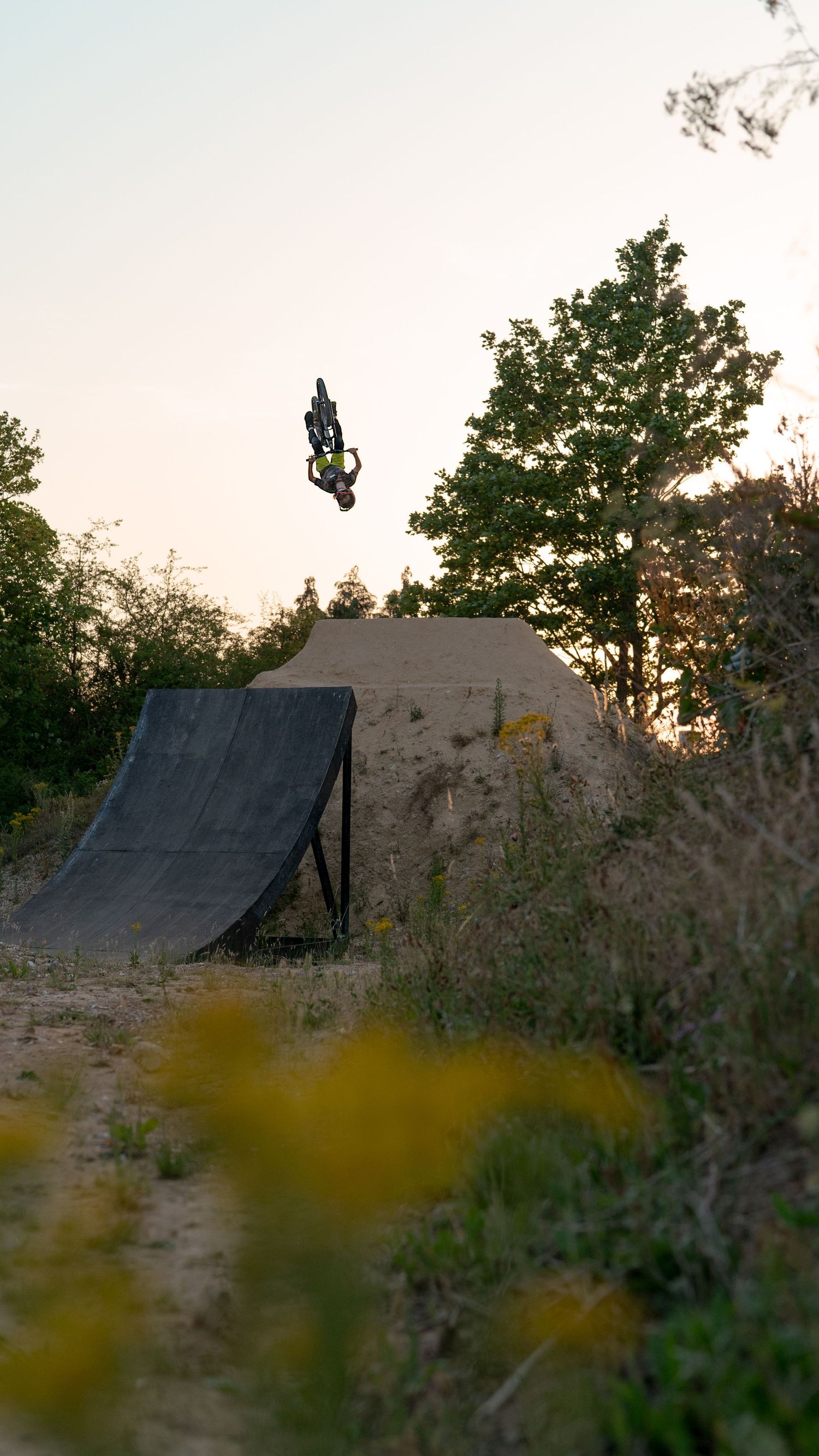 BMX rider performing a high aerial trick off a dirt ramp at sunset, surrounded by trees and natural terrain.