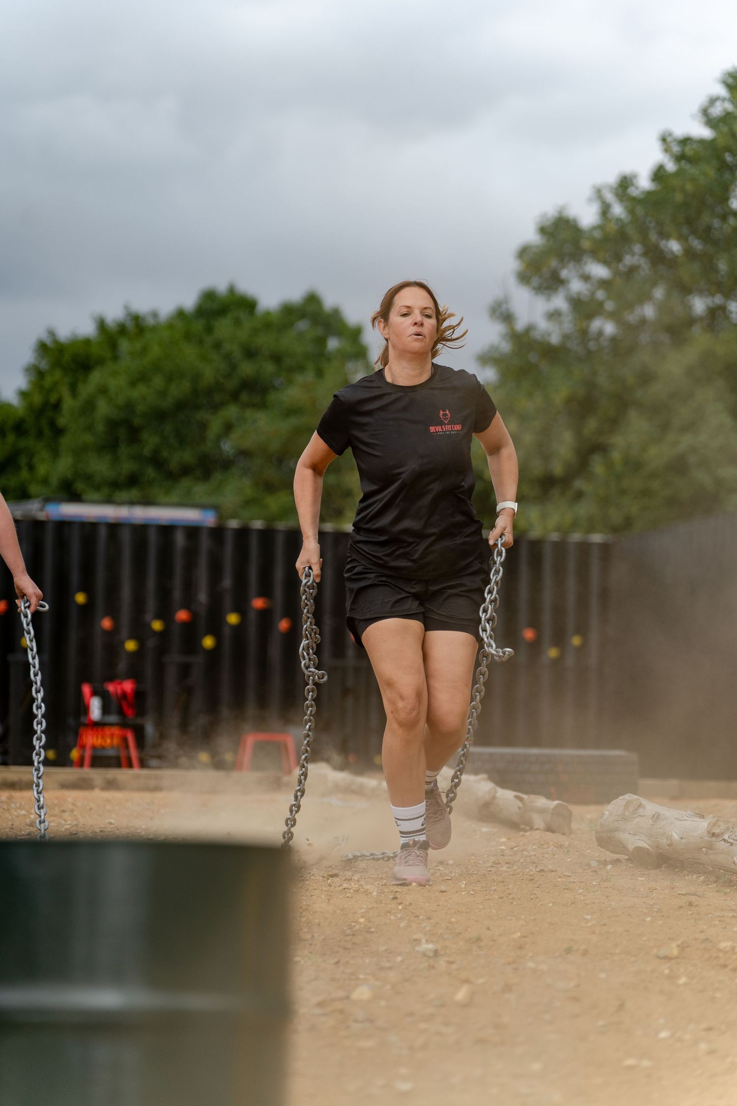 Woman in black shirt drags heavy chains across sandy obstacle course during outdoor fitness competition.