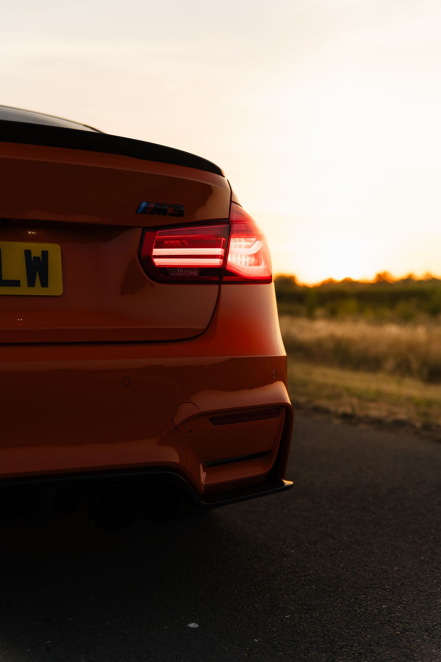 Rear view of an orange BMW M3 at sunset, with glowing red tail lights and a UK license plate on a country road.