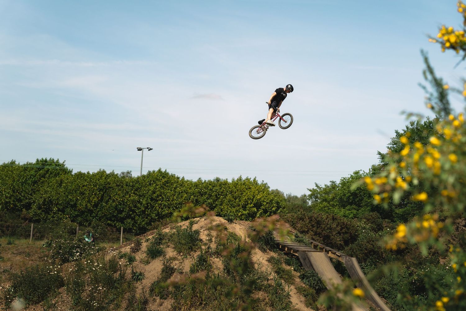BMX rider performing a high aerial trick jump over a dirt mound at an outdoor bike park with green bushes and blue sky.