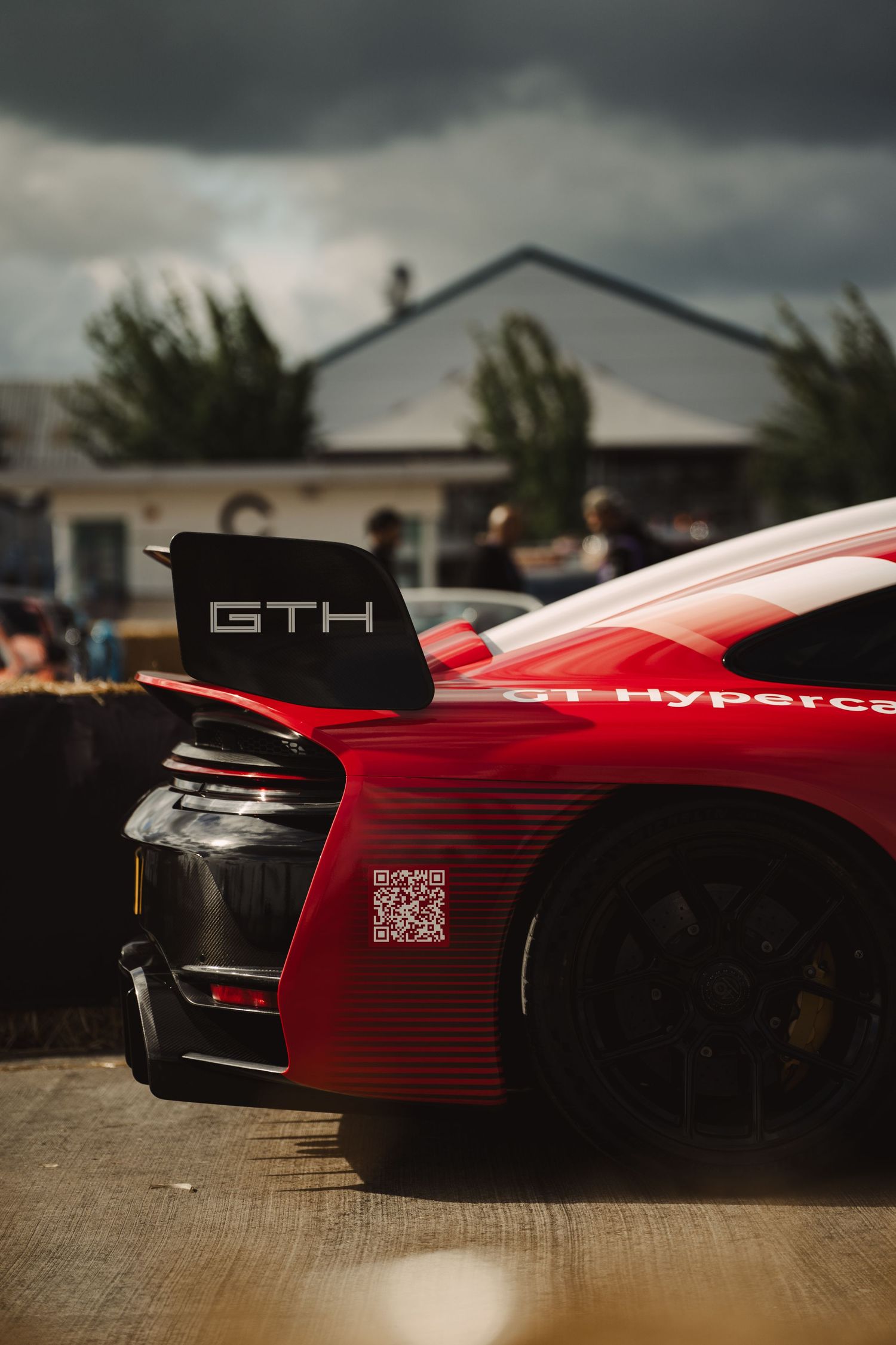 Red GTH hypercar rear spoiler with QR code on display at an outdoor automotive event under cloudy skies.