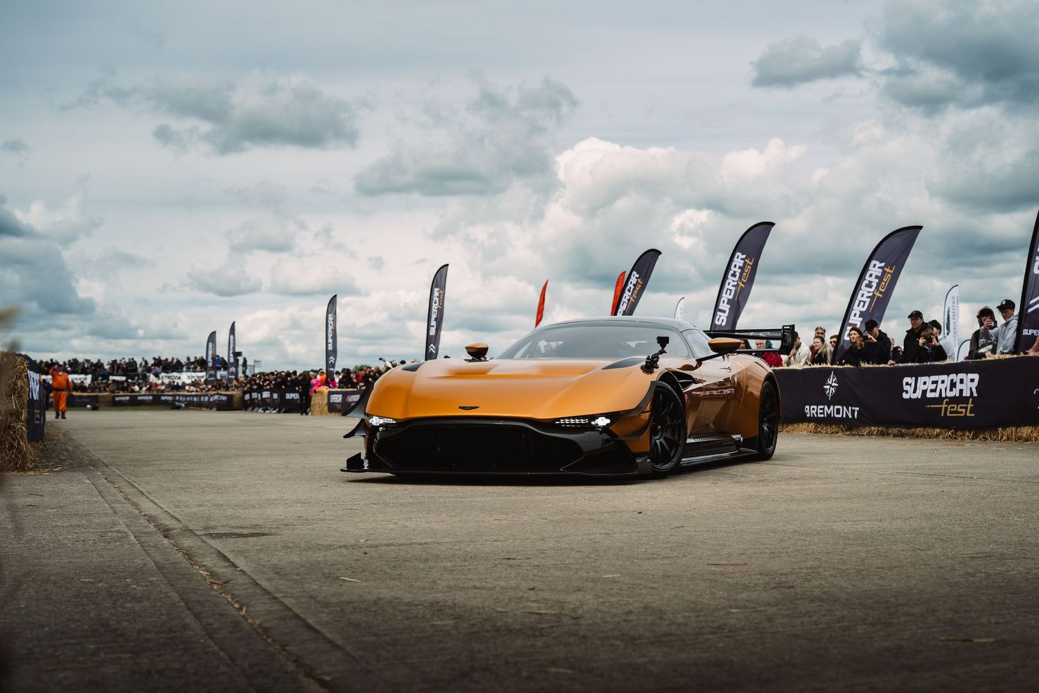 Orange Aston Martin Vulcan supercar on display at an outdoor automotive event with colorful flags and cloudy sky.