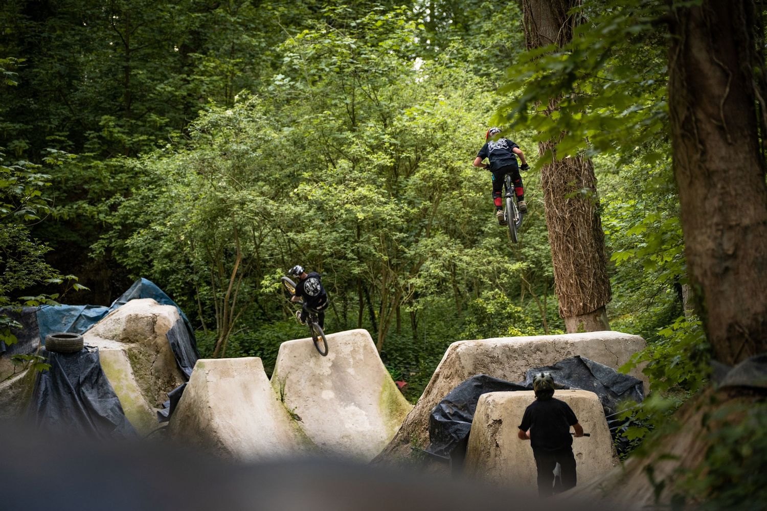 Mountain bikers performing jumps over large concrete blocks in a lush forest setting.