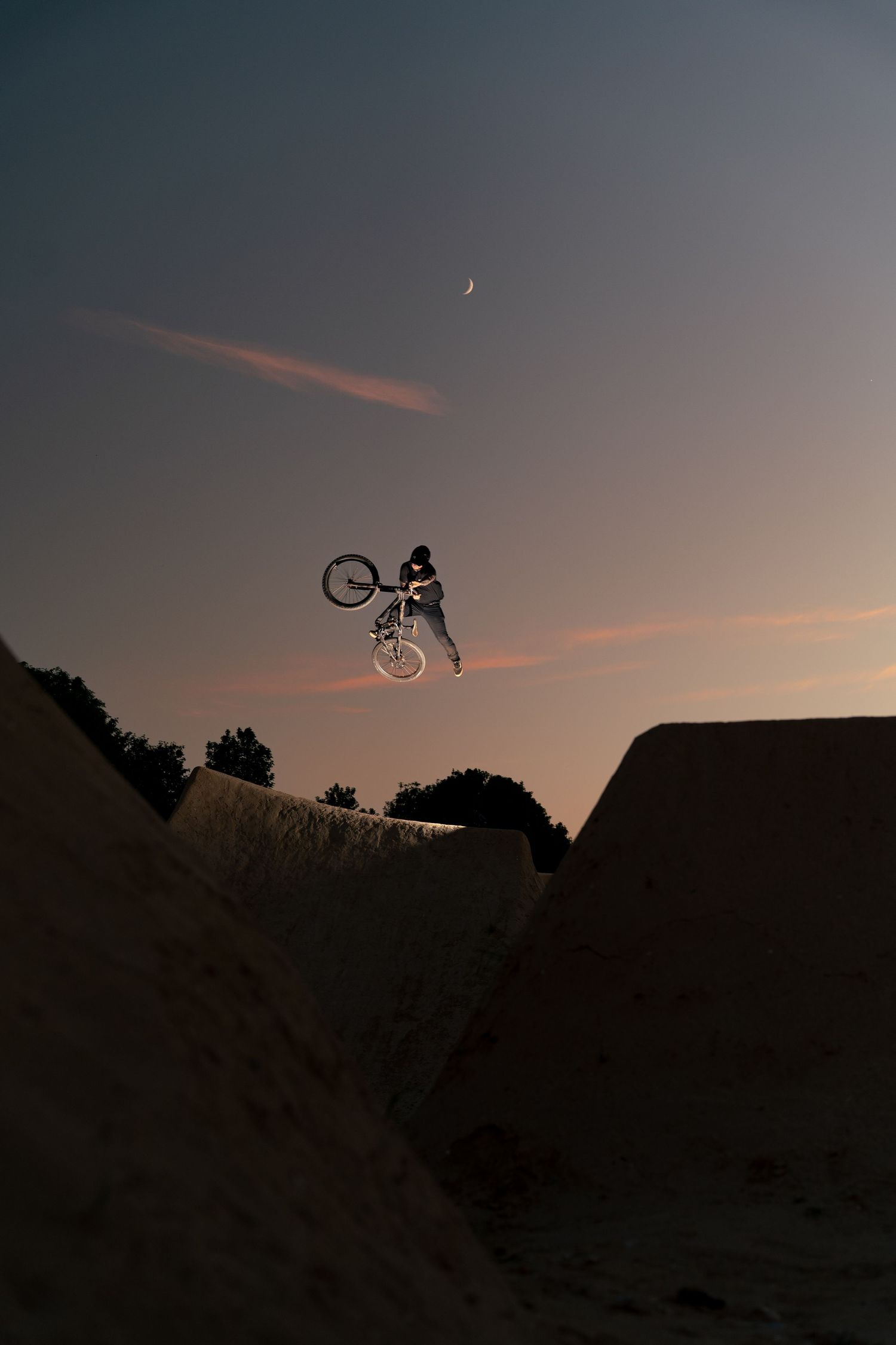 Silhouette of BMX rider performing aerial trick at sunset with crescent moon visible in dramatic twilight sky.