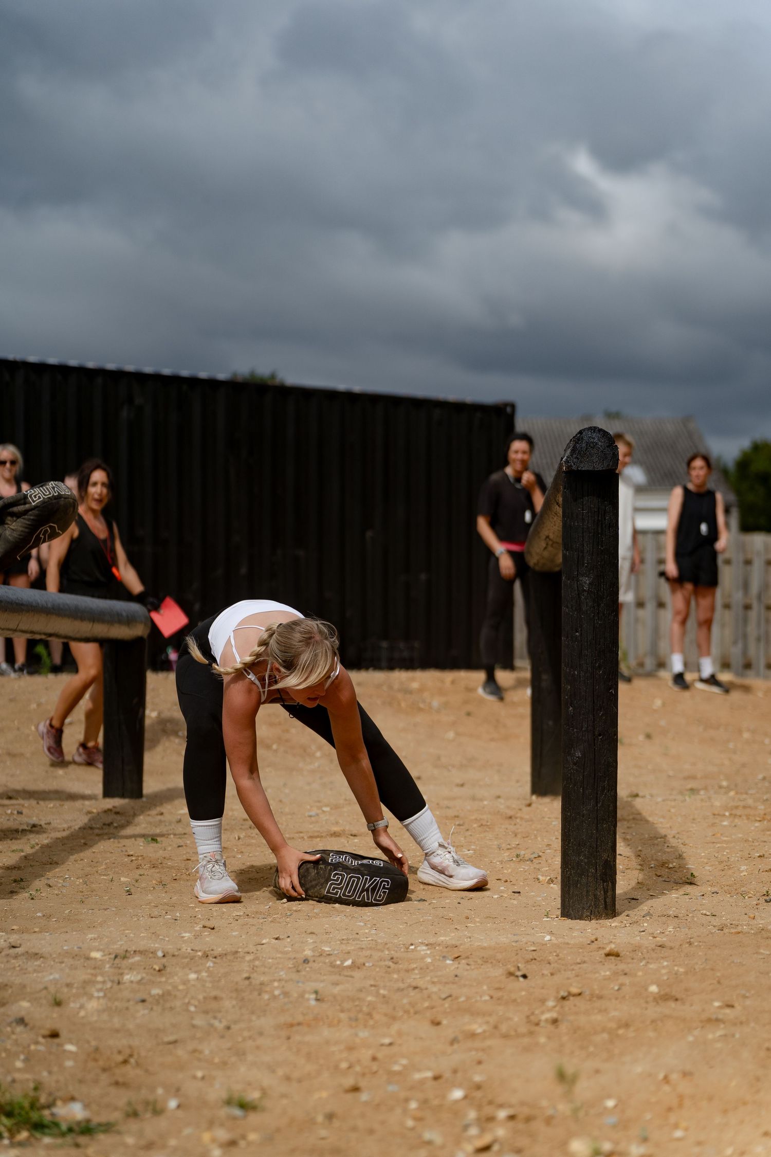Woman bending down to pick up a weighted sandbag during an outdoor fitness obstacle course challenge.