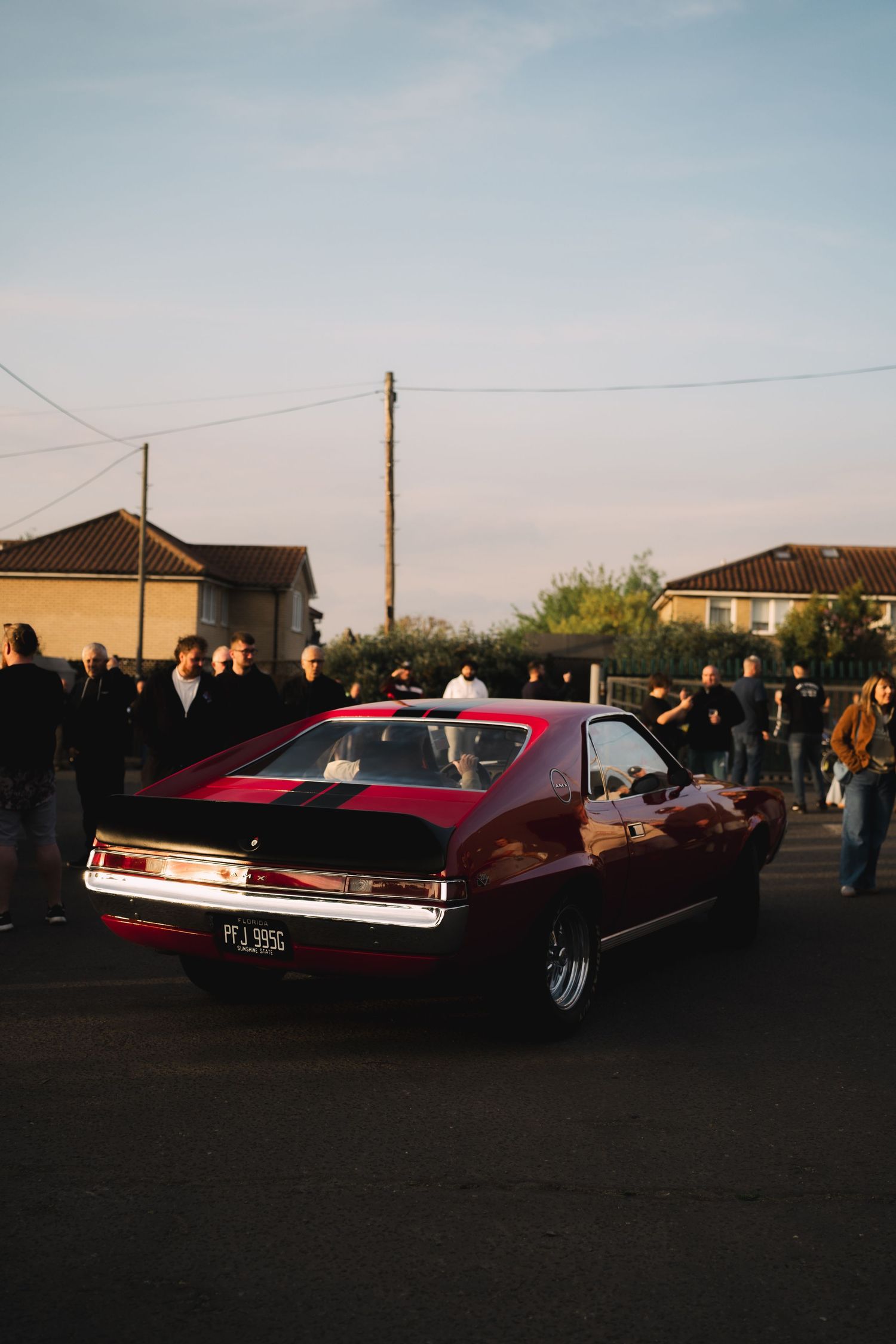 A classic red muscle car with black racing stripes parked on a street surrounded by spectators at golden hour.