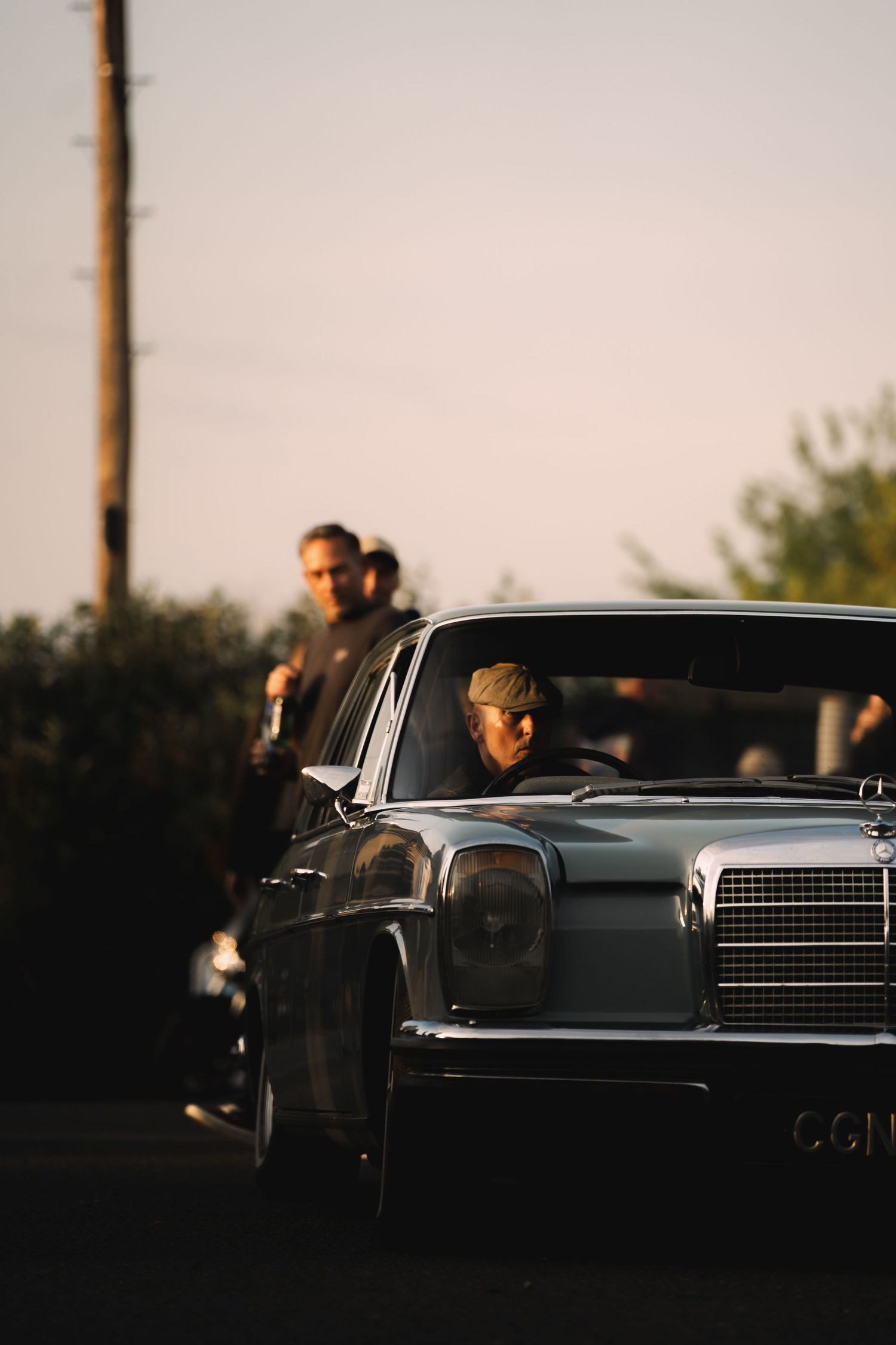 Vintage Mercedes-Benz sedan parked at golden hour with people standing in background near trees.