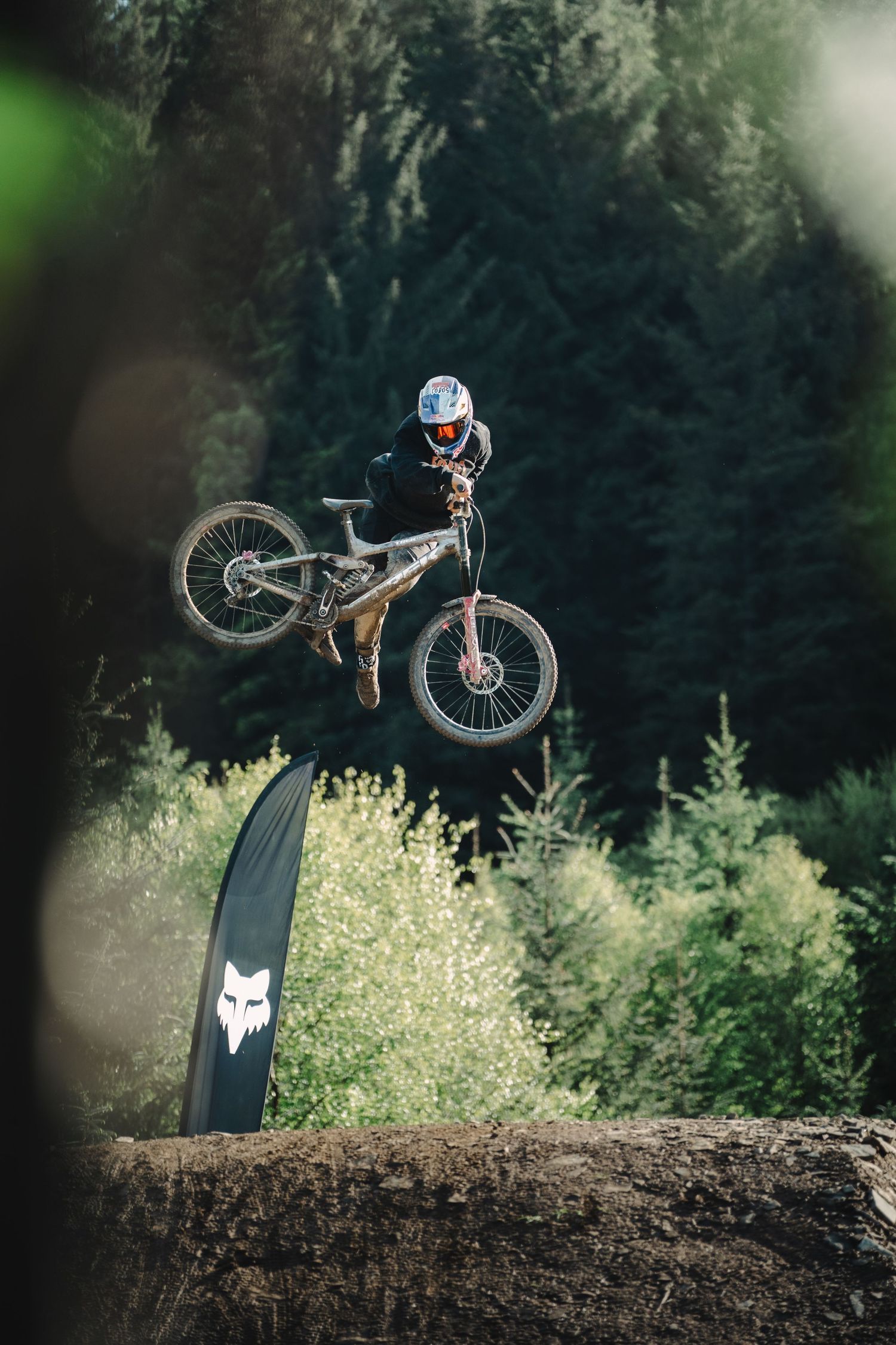 Mountain biker performs aerial trick over dirt jump at Fox-sponsored downhill event, surrounded by dense forest backdrop.