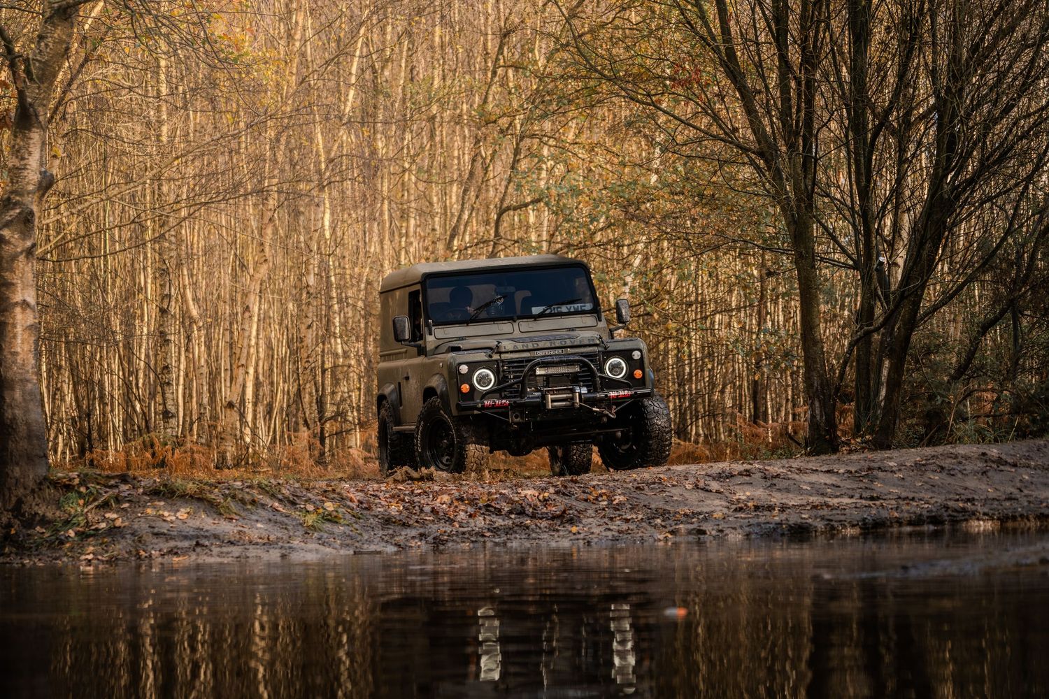 Dark Land Rover Defender reflected in a still forest puddle surrounded by golden autumn trees.