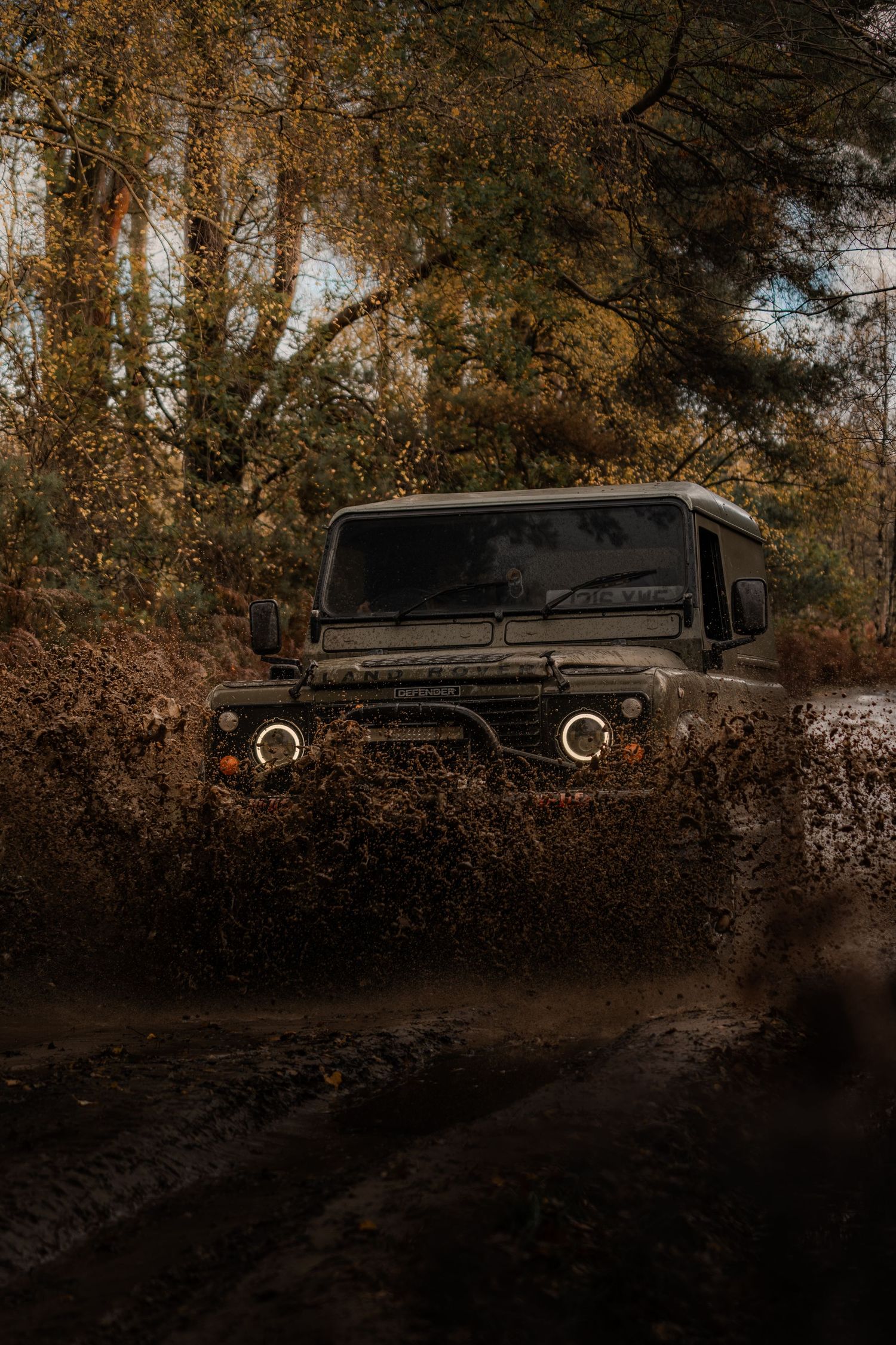 Land Rover Defender splashing through a deep muddy puddle on an autumn forest off-road trail.