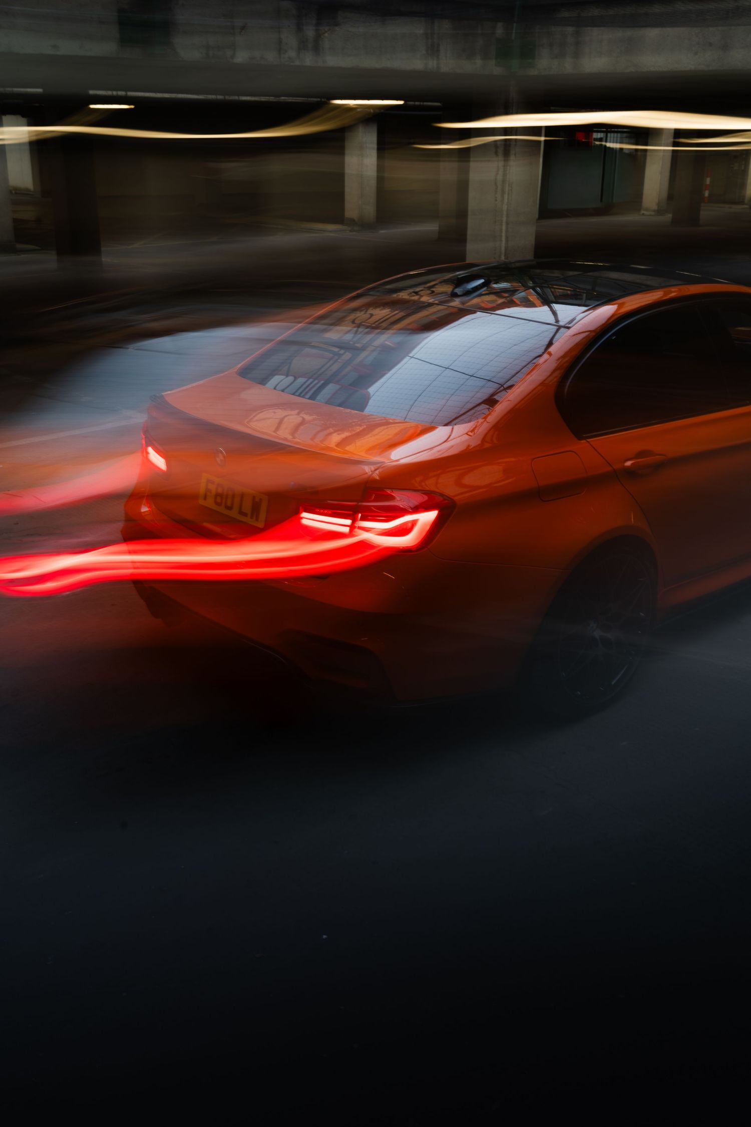 Orange BMW sedan speeding through dark parking garage with motion blur and glowing red taillights.