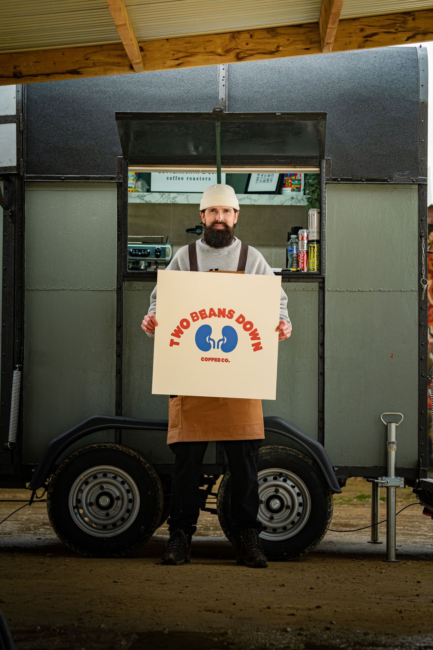 Bearded barista holds Two Beans Down Coffee Co. sign outside a mobile coffee trailer.