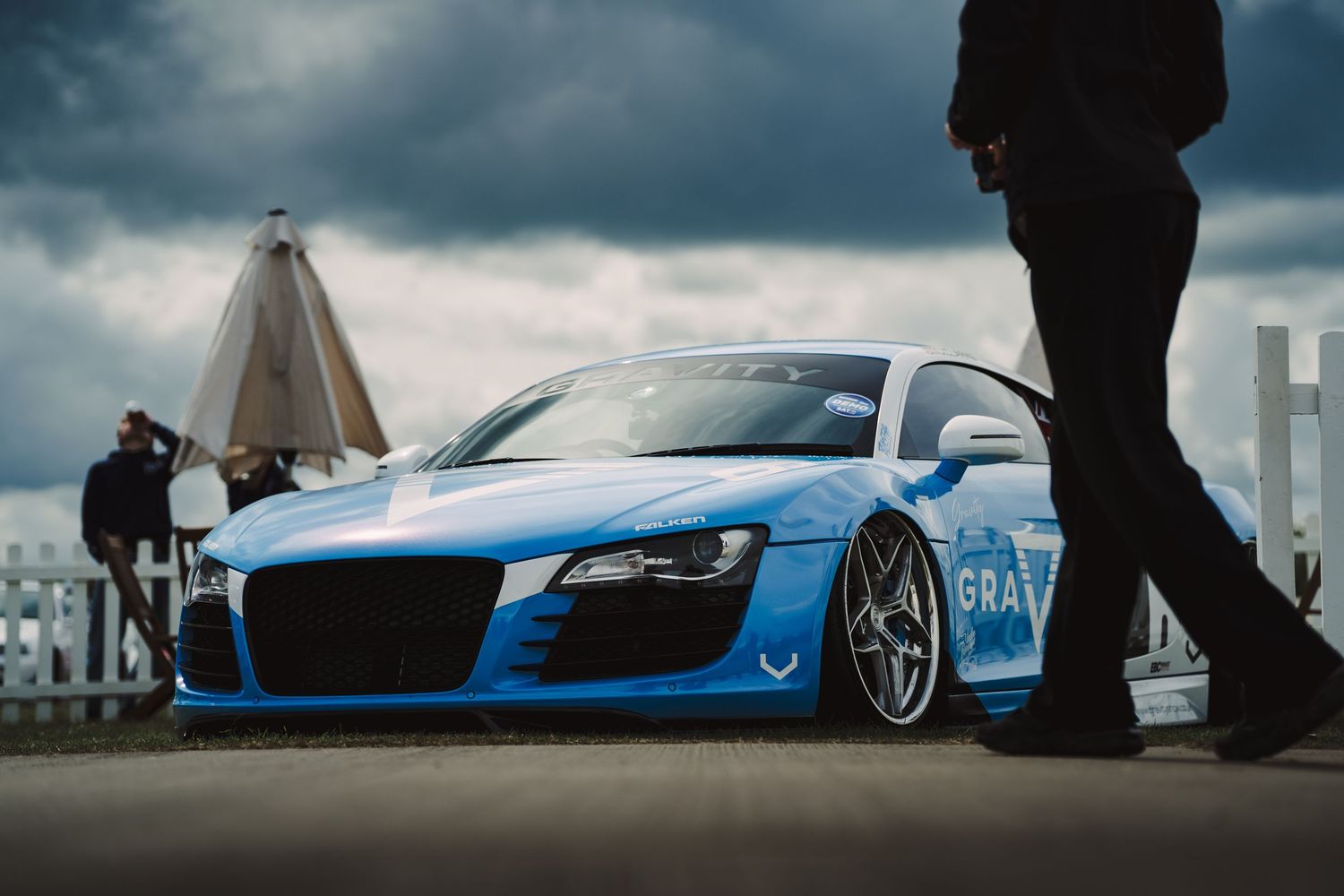 A lowered blue Audi R8 supercar on custom wheels displayed outdoors under dramatic cloudy skies at a car show.