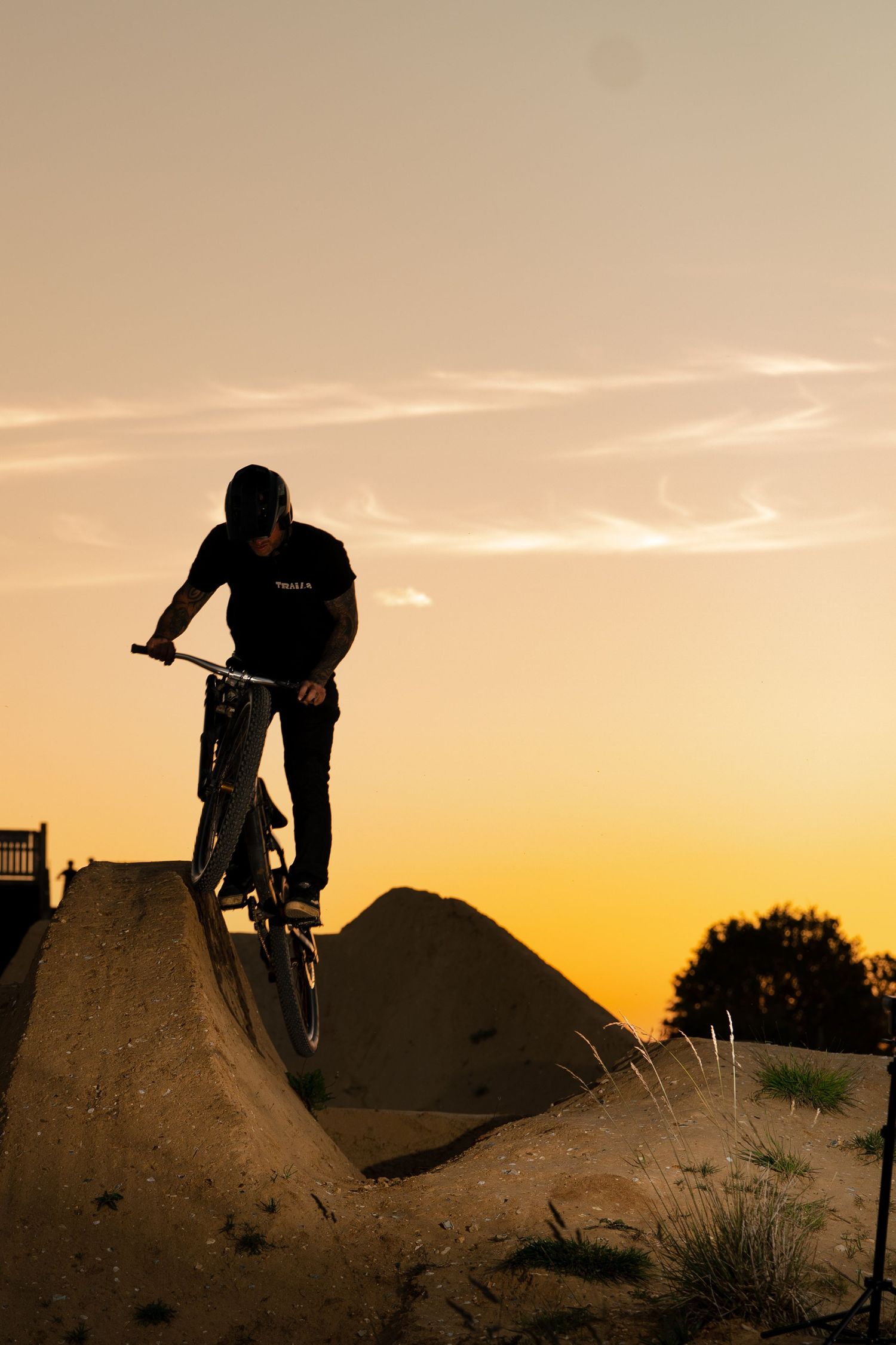 BMX rider silhouette descending a dirt ramp at a skate park during golden hour sunset.