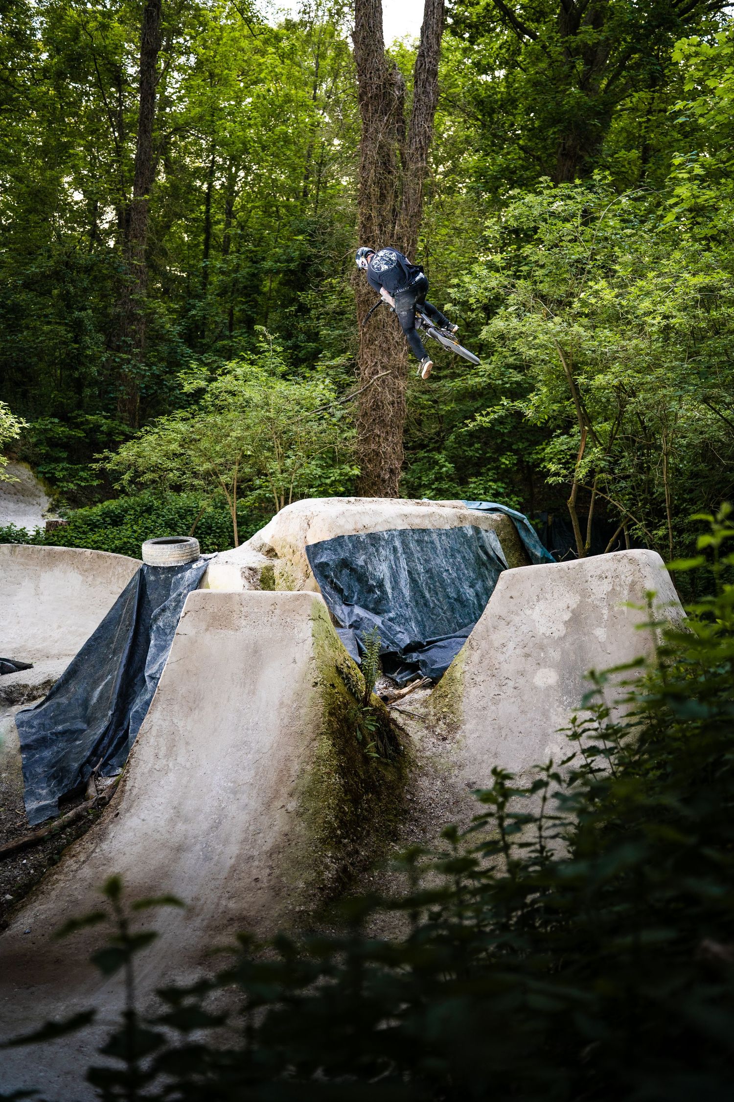 BMX rider performing aerial trick over concrete skate park ramps surrounded by lush green forest trees.