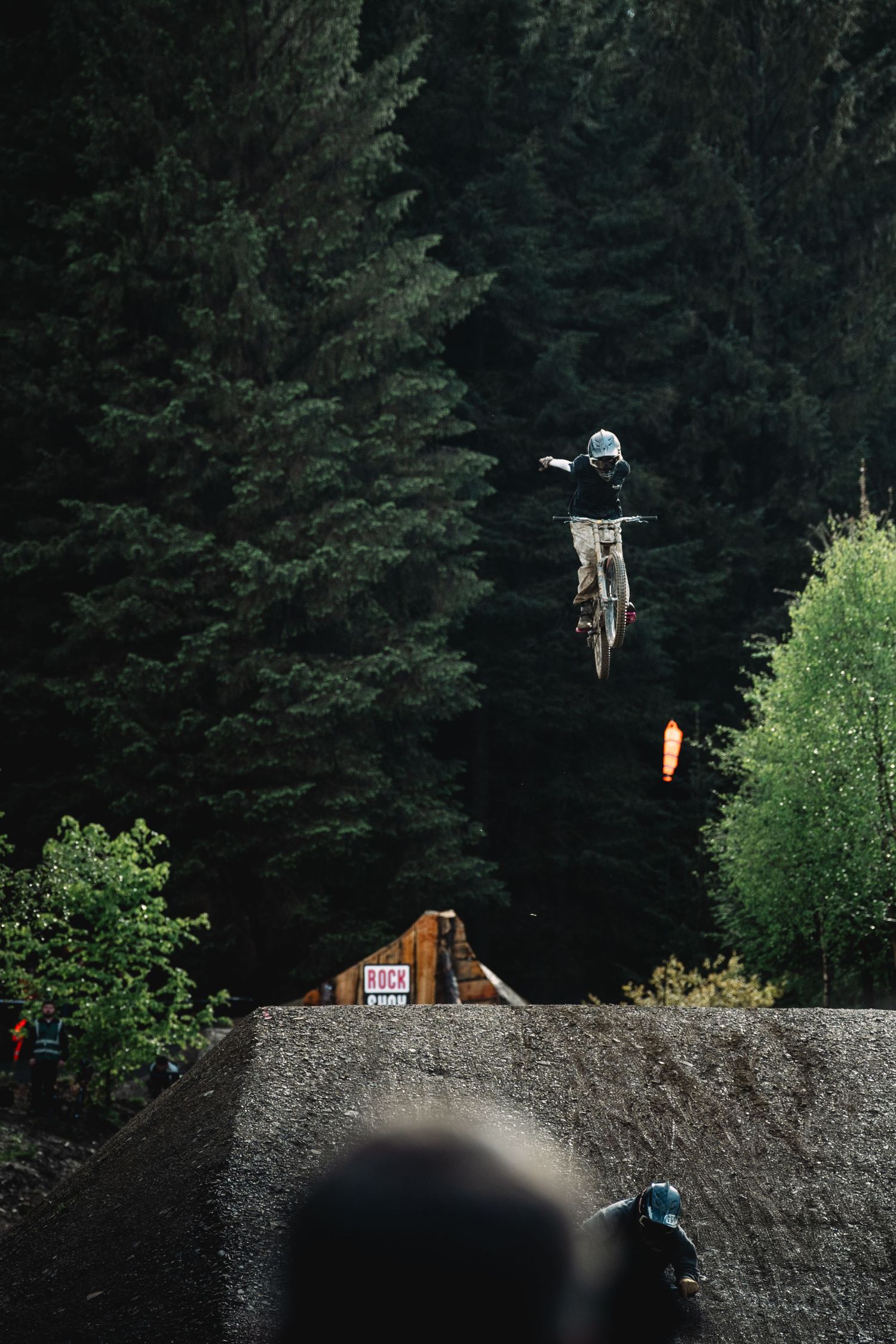 A mountain biker performs a high aerial jump over a dirt ramp surrounded by dense evergreen forest.