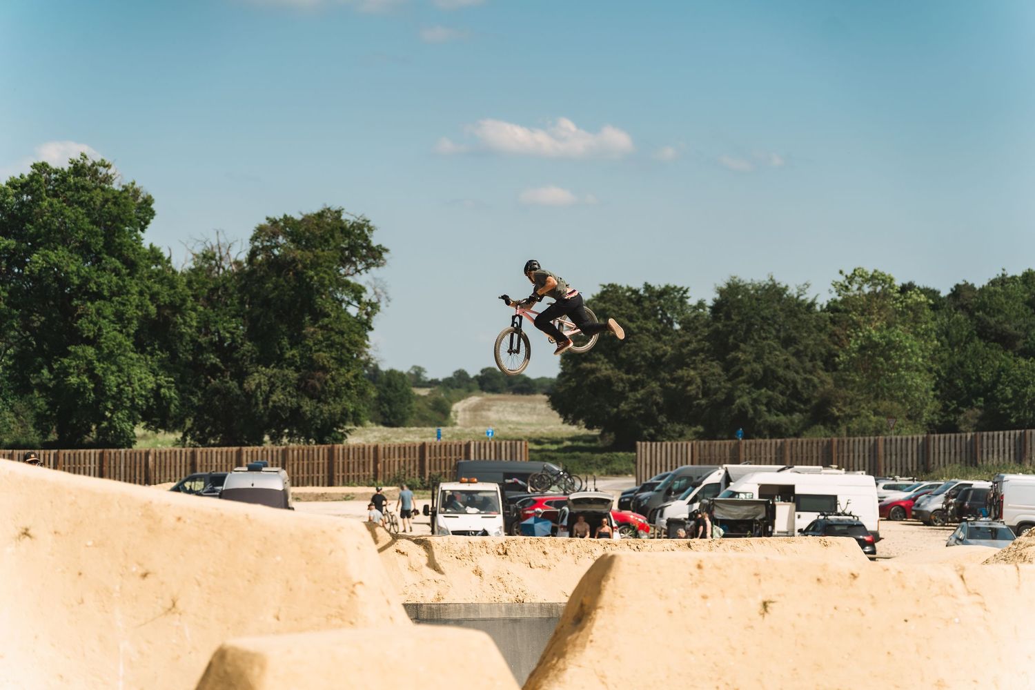 BMX rider performs high aerial trick over dirt jumps at outdoor bike park on a sunny day.