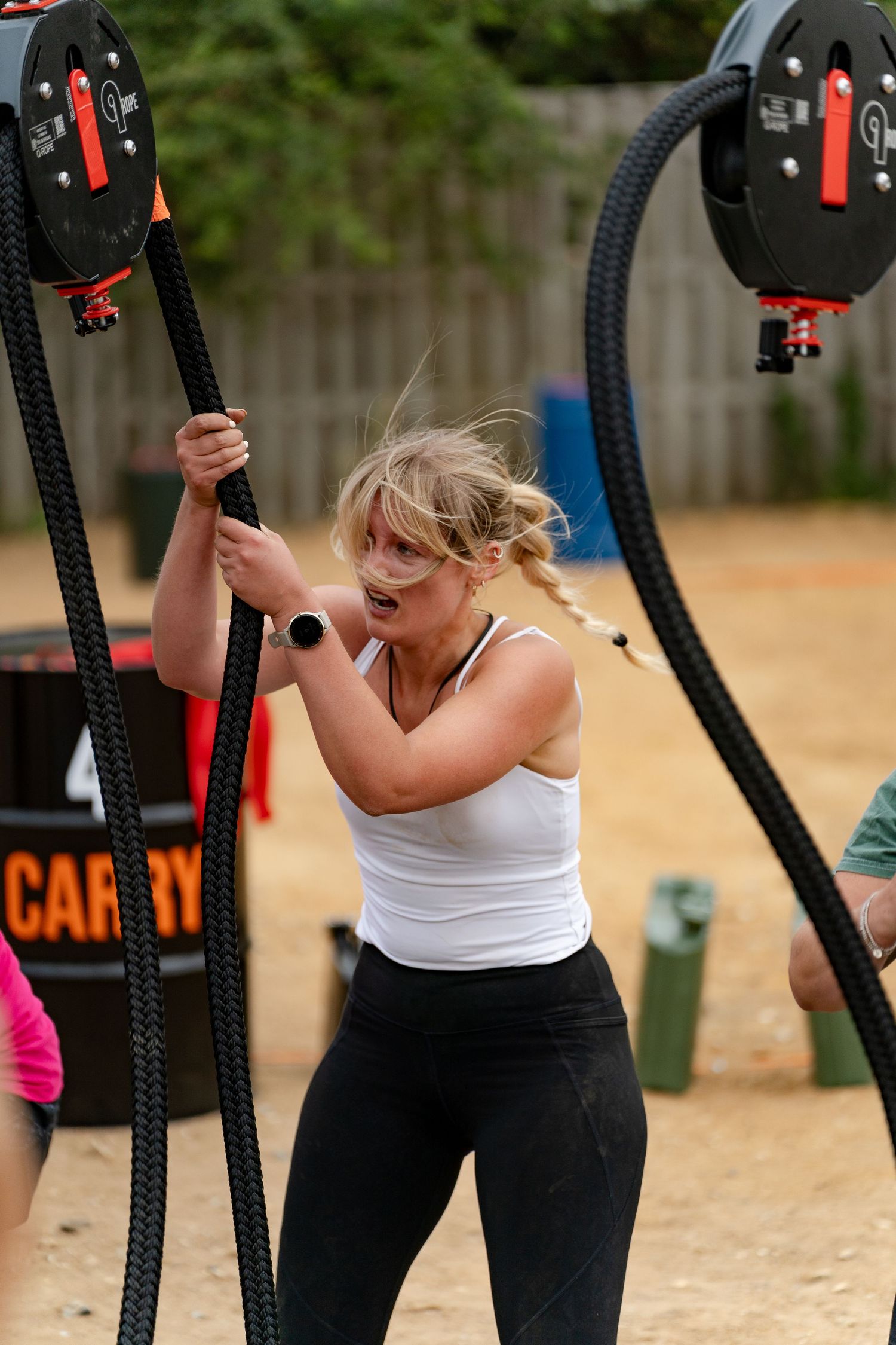 Blonde woman in white tank top pulling heavy battle ropes during outdoor fitness training competition.