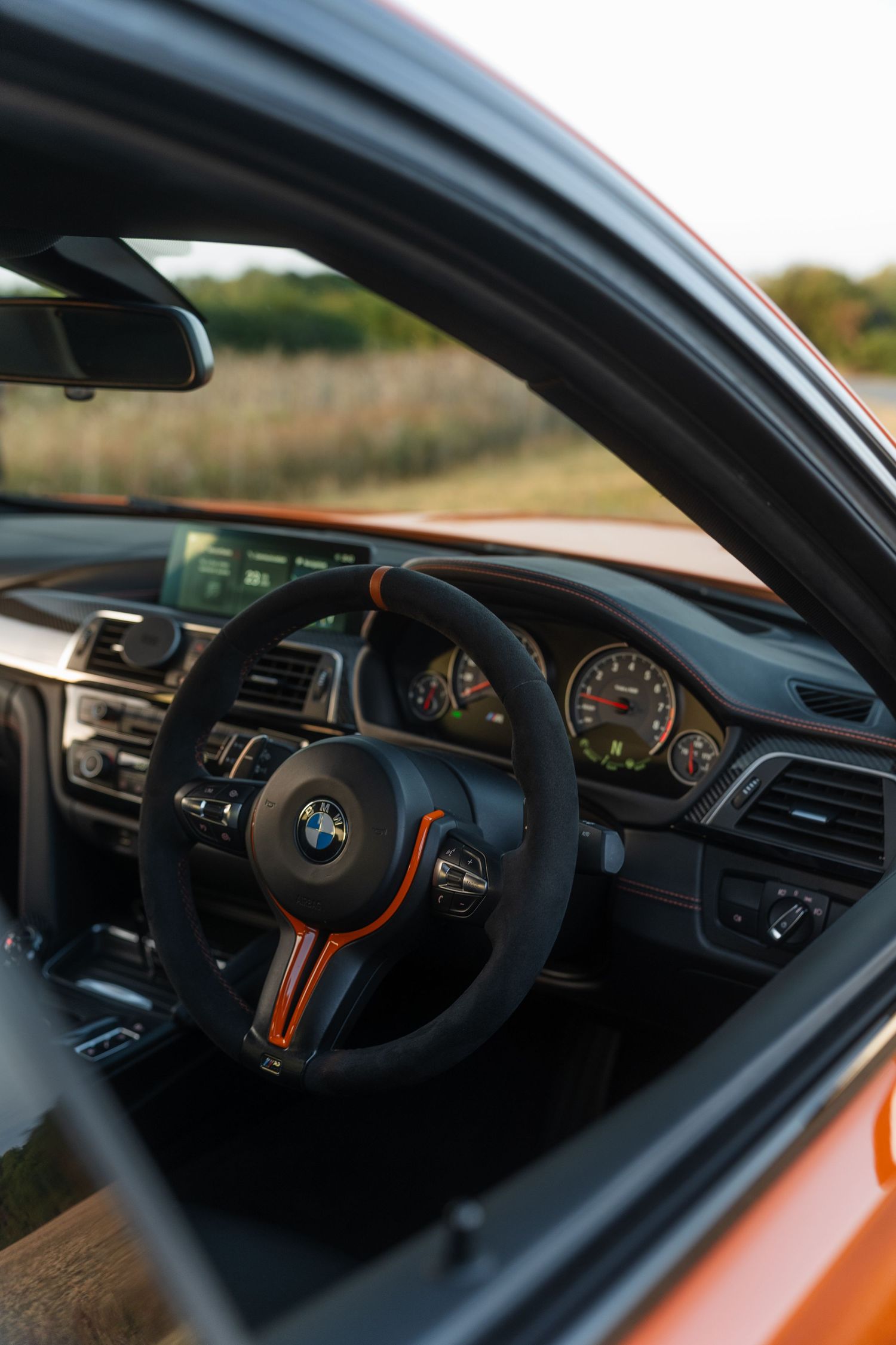 BMW M4 interior featuring a sport steering wheel with orange accents, analog gauges, and carbon trim in an orange exterior car.