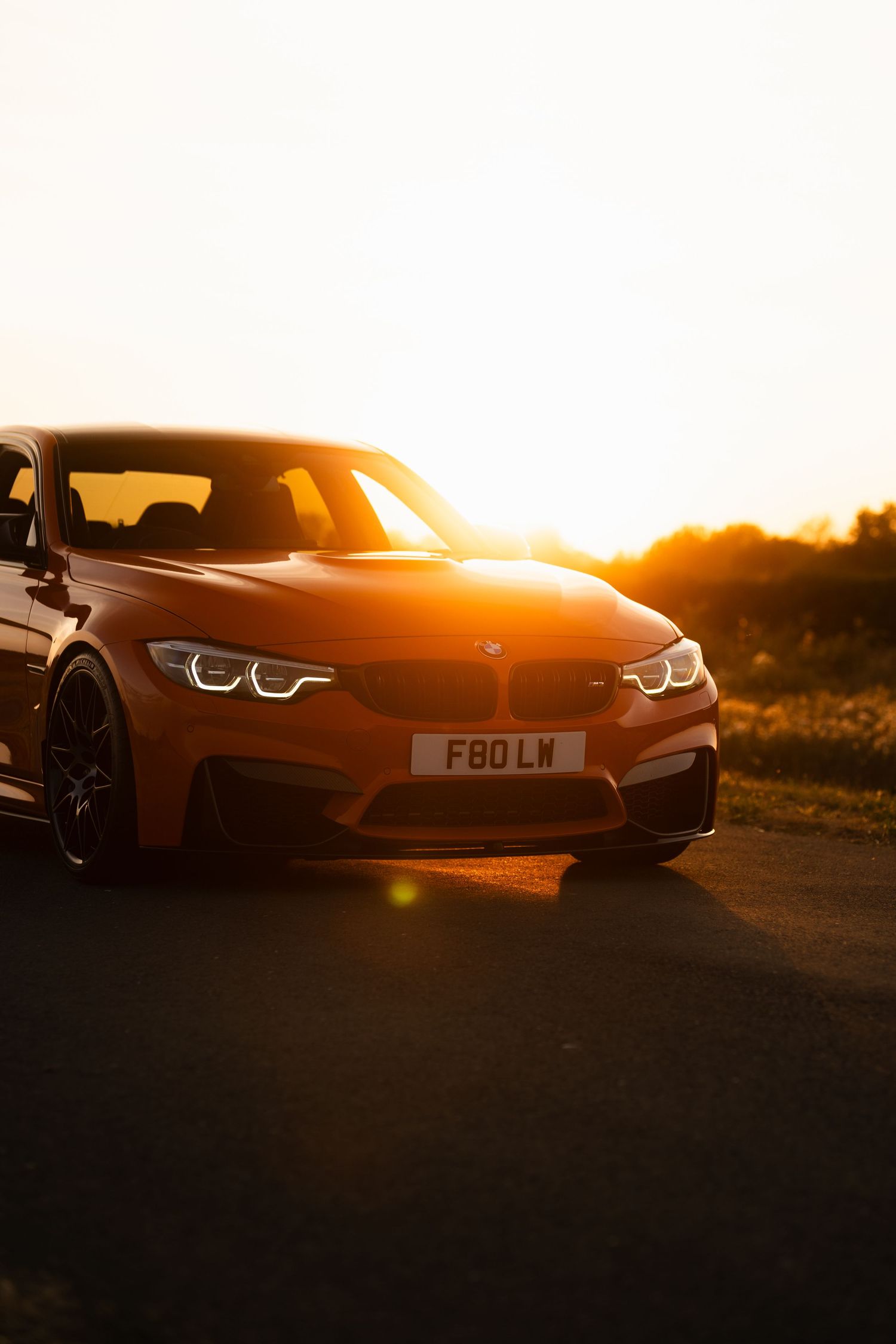 Orange BMW M3 F80 photographed at golden sunset on an empty road, with warm sunlight reflecting off the hood.