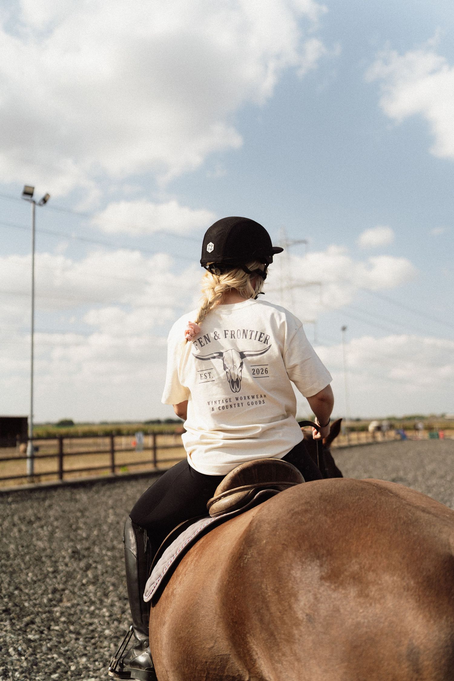 Young equestrian rider in white graphic tee and black helmet riding a brown horse in an outdoor arena.