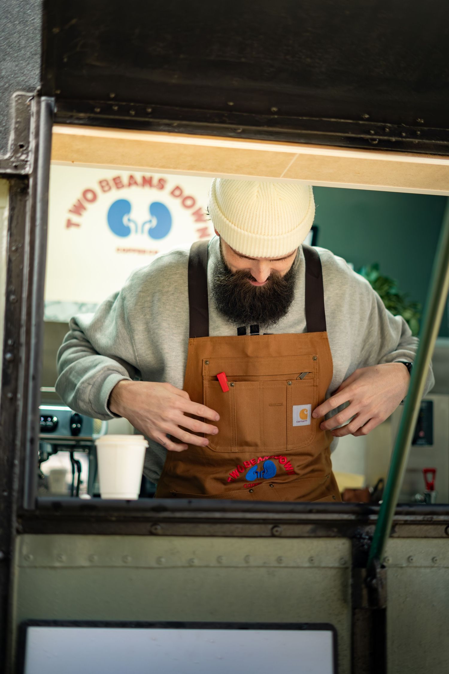 Bearded barista in white beanie adjusting brown Carhartt apron inside Two Beans Down coffee truck.