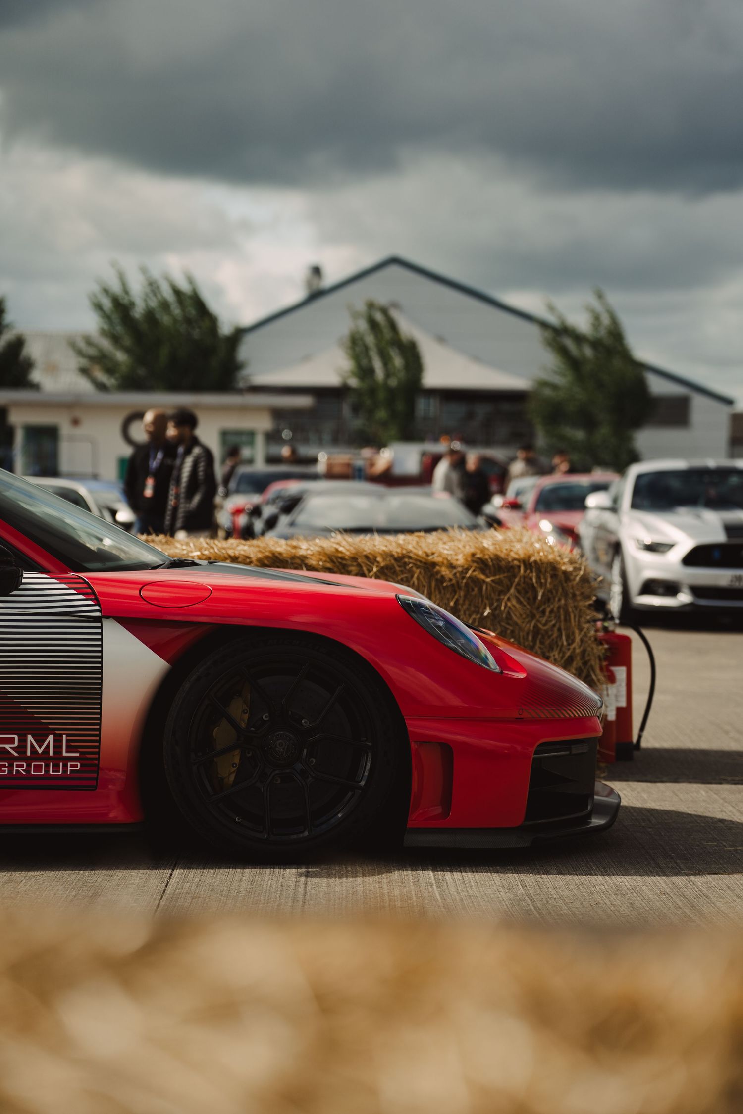 Red RML Group Porsche 911 GT2 RS race car at a motorsport event with hay bales and Ford Mustang in background.