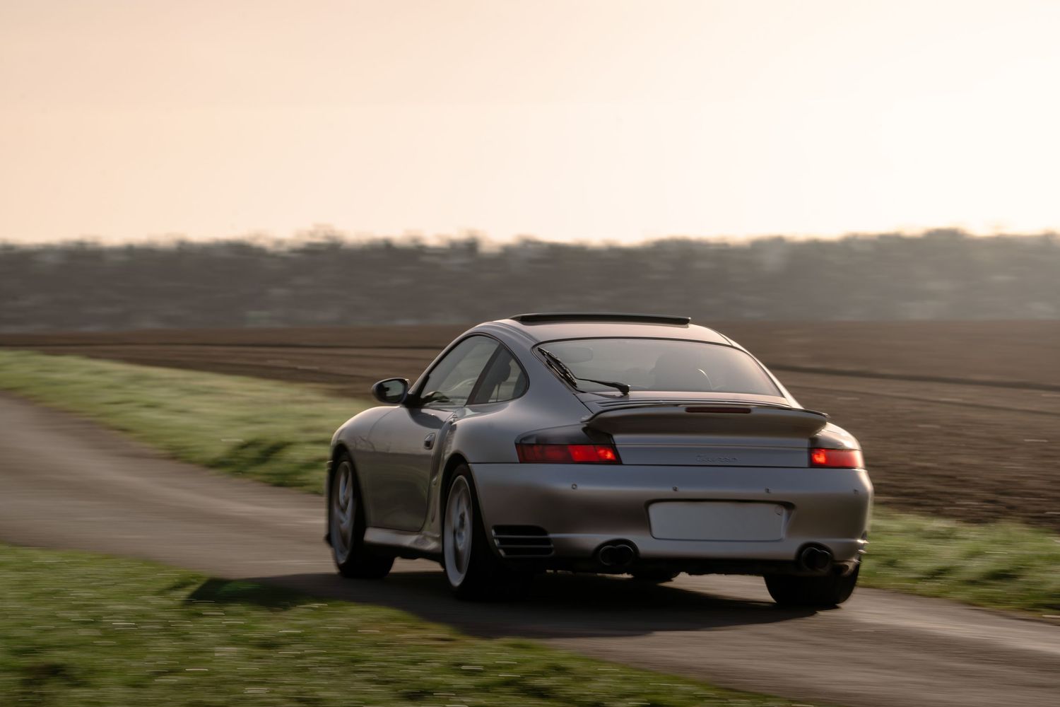 Silver Porsche 911 Turbo rear view speeding along a rural road at golden hour sunset.