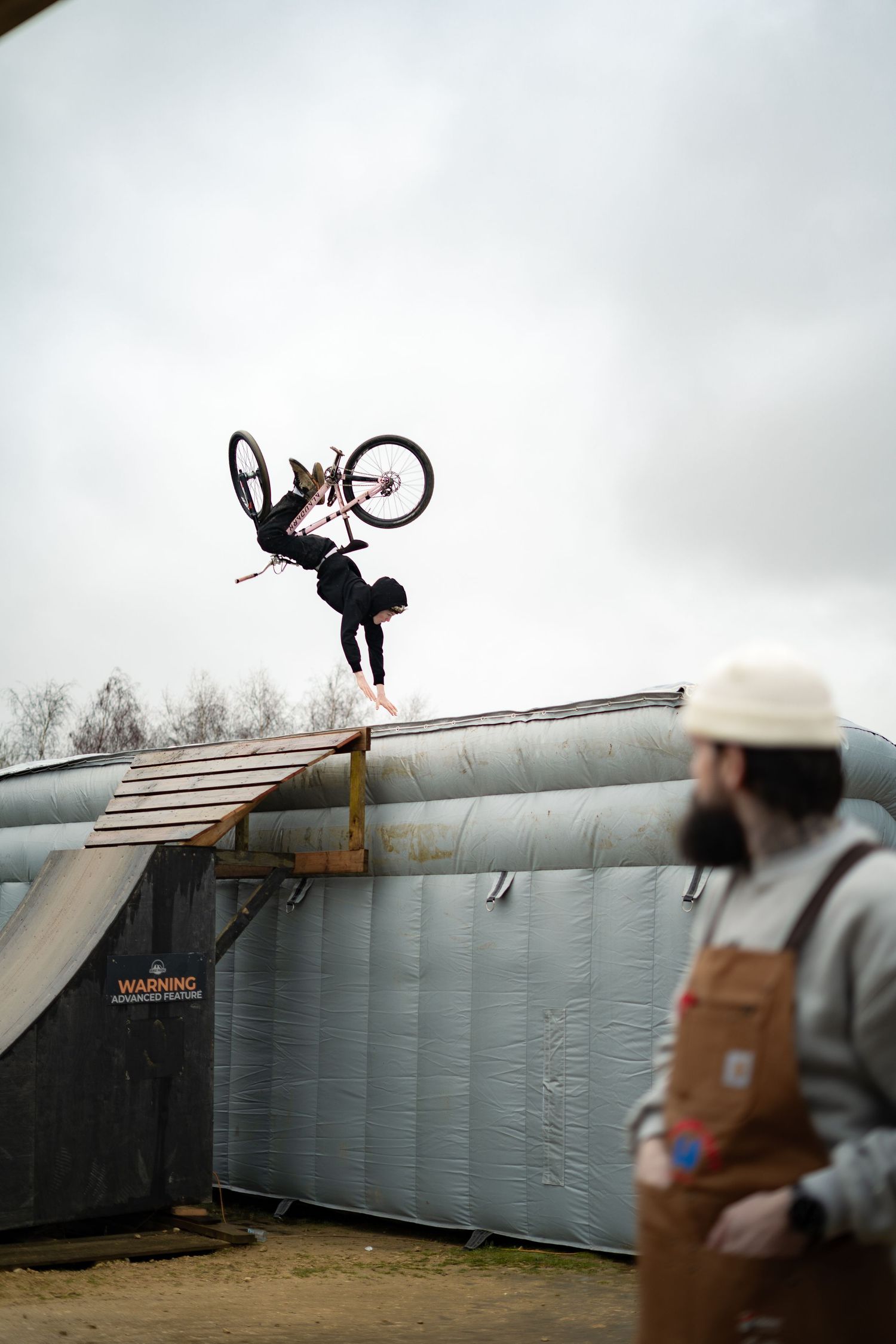 A BMX rider executes a dramatic backflip over a ramp while a bearded man in an apron observes.