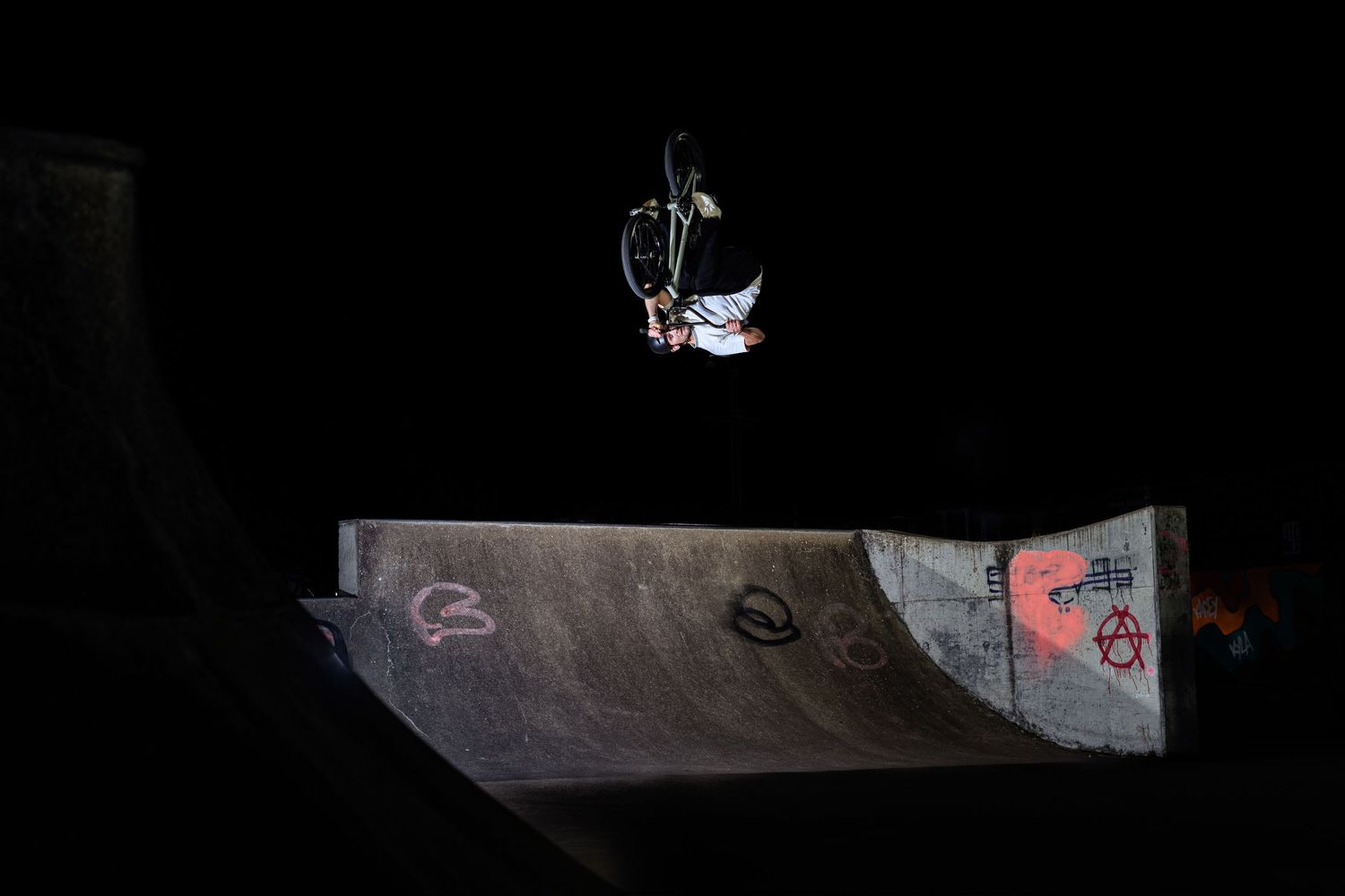 BMX rider performing aerial trick on halfpipe ramp at night skatepark with graffiti walls.