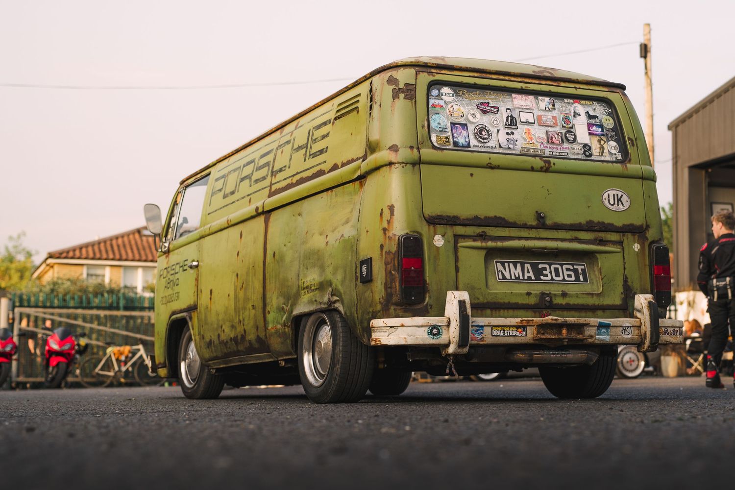 Vintage green Volkswagen T2 van with Porsche branding and stickers, lowered stance, parked on pavement at an outdoor event.