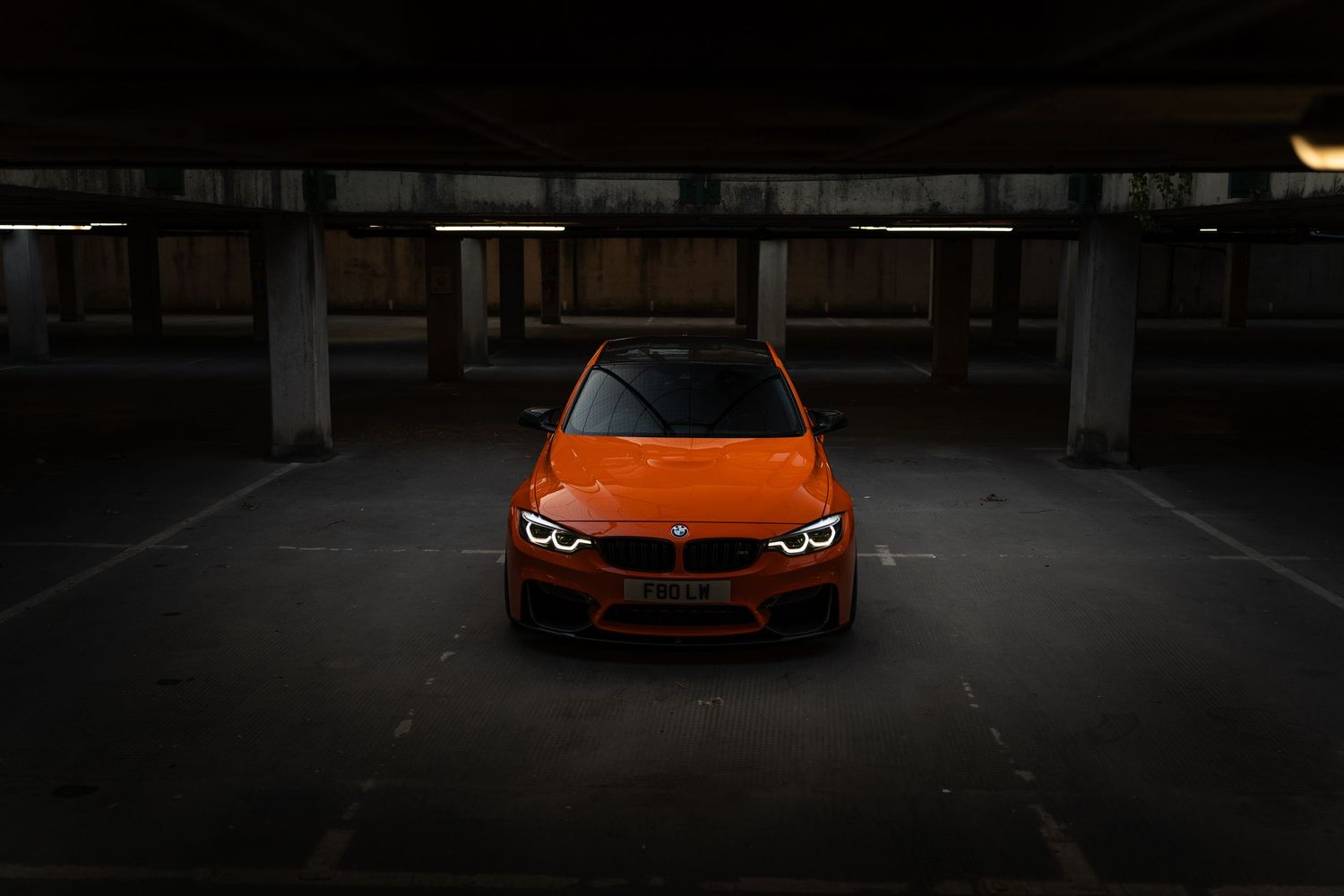 Vibrant orange BMW M3 parked in a dark parking garage, showcasing its aggressive front fascia and LED headlights.