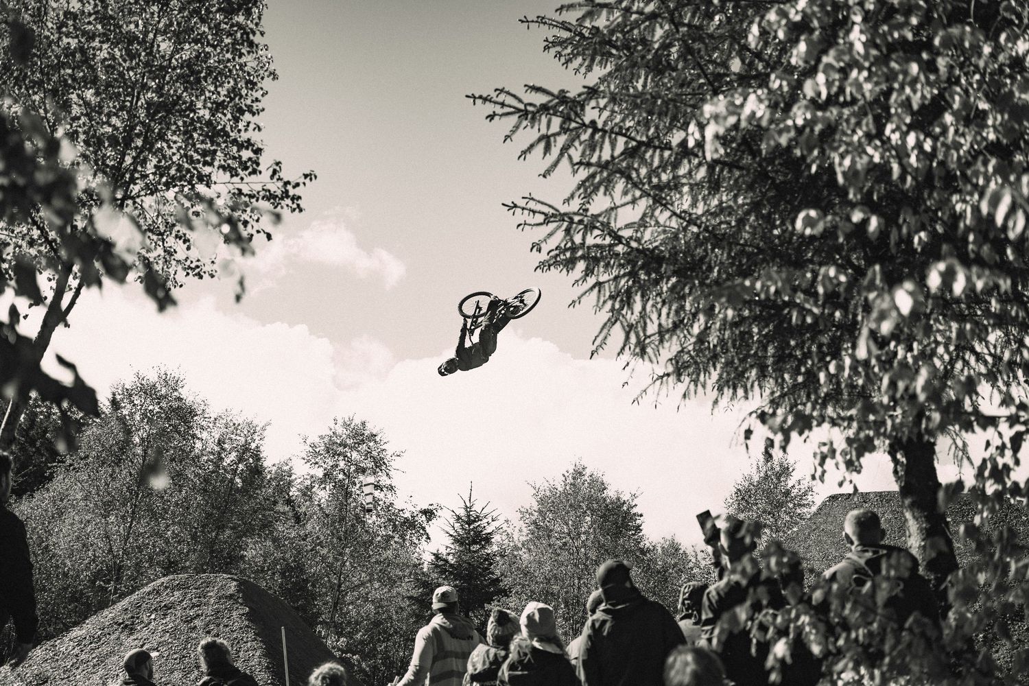 Black and white photo of a BMX rider performing a high jump trick over a crowd of spectators surrounded by trees.