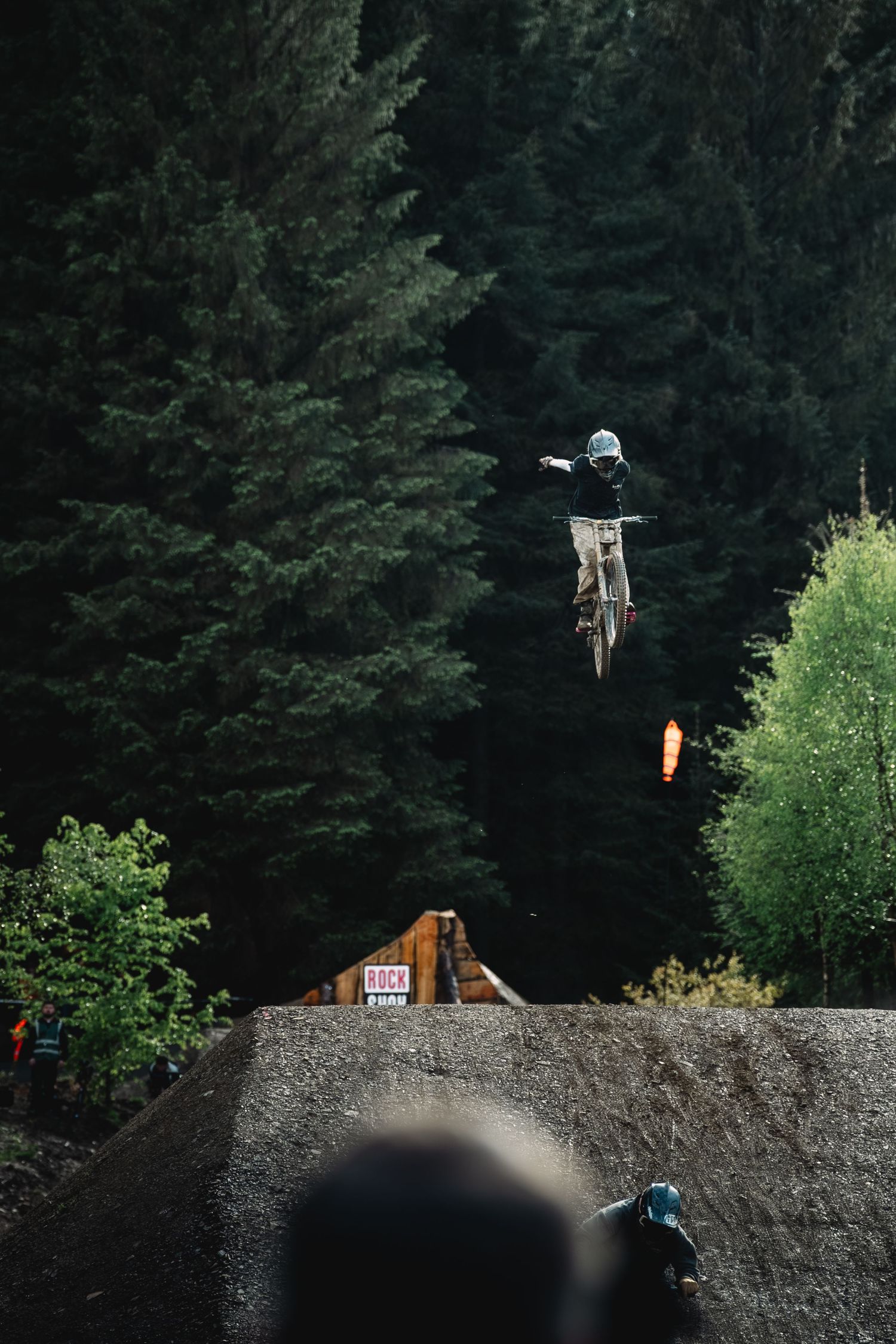 A mountain biker performs a dramatic aerial jump over a wooden ramp with dense evergreen forest in the background.