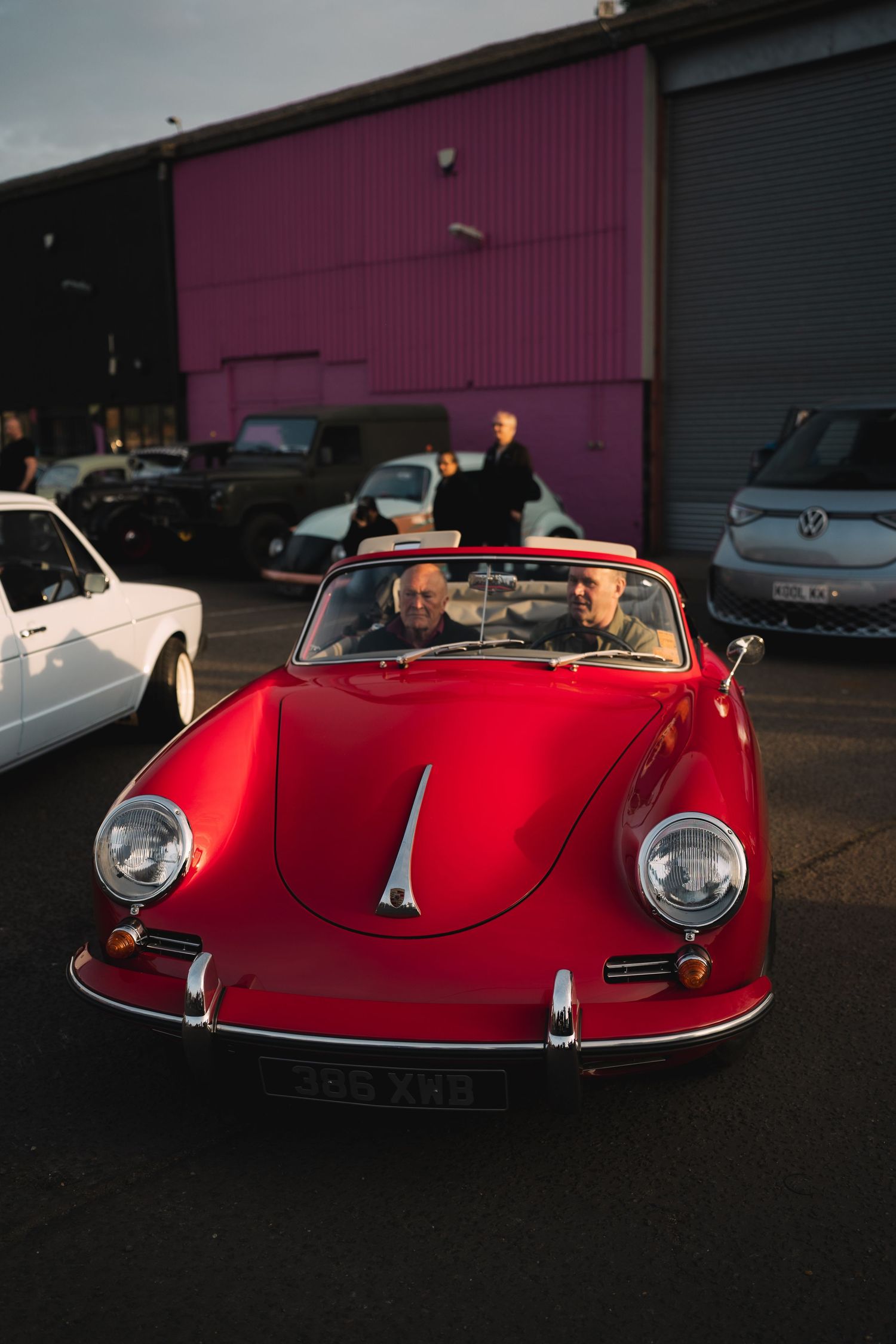 Two men ride in a classic red Porsche 356 convertible parked outside an industrial building at dusk.