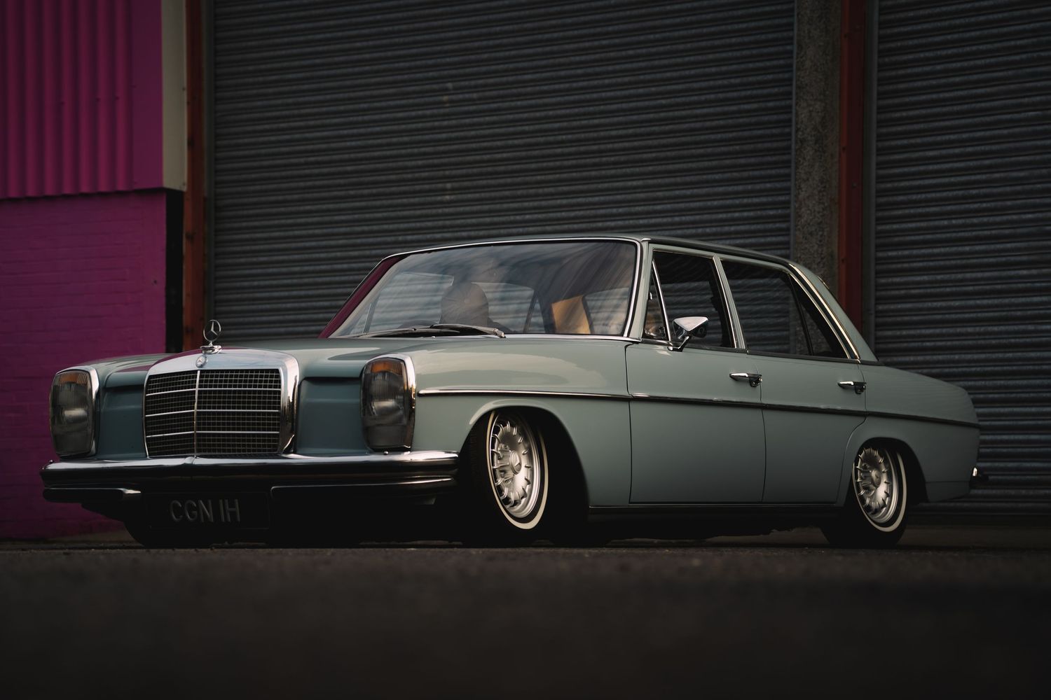 Vintage Mercedes-Benz W114 sedan in light blue, lowered stance, parked against industrial garage doors at night.