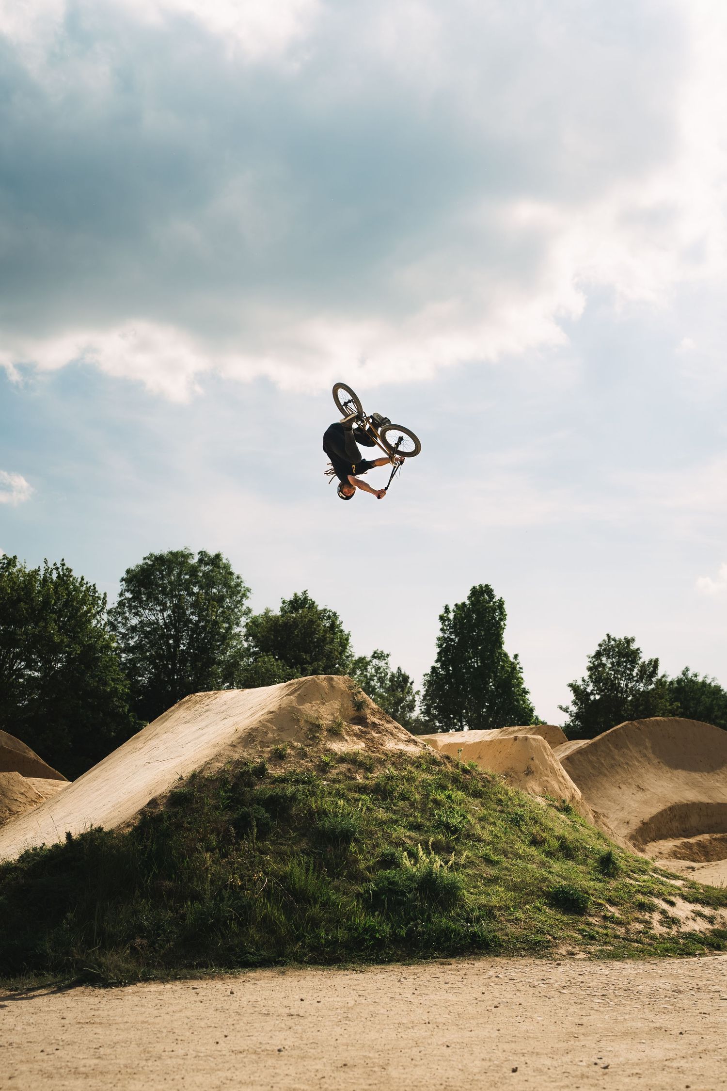 BMX rider executes an aerial trick over dirt jumps at an outdoor bike park under cloudy skies.