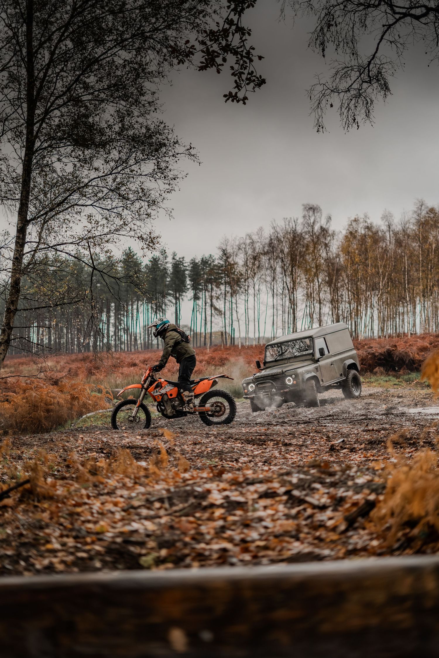 Motocross rider on orange dirt bike races alongside a dark green Land Rover Defender on a muddy forest trail in autumn.